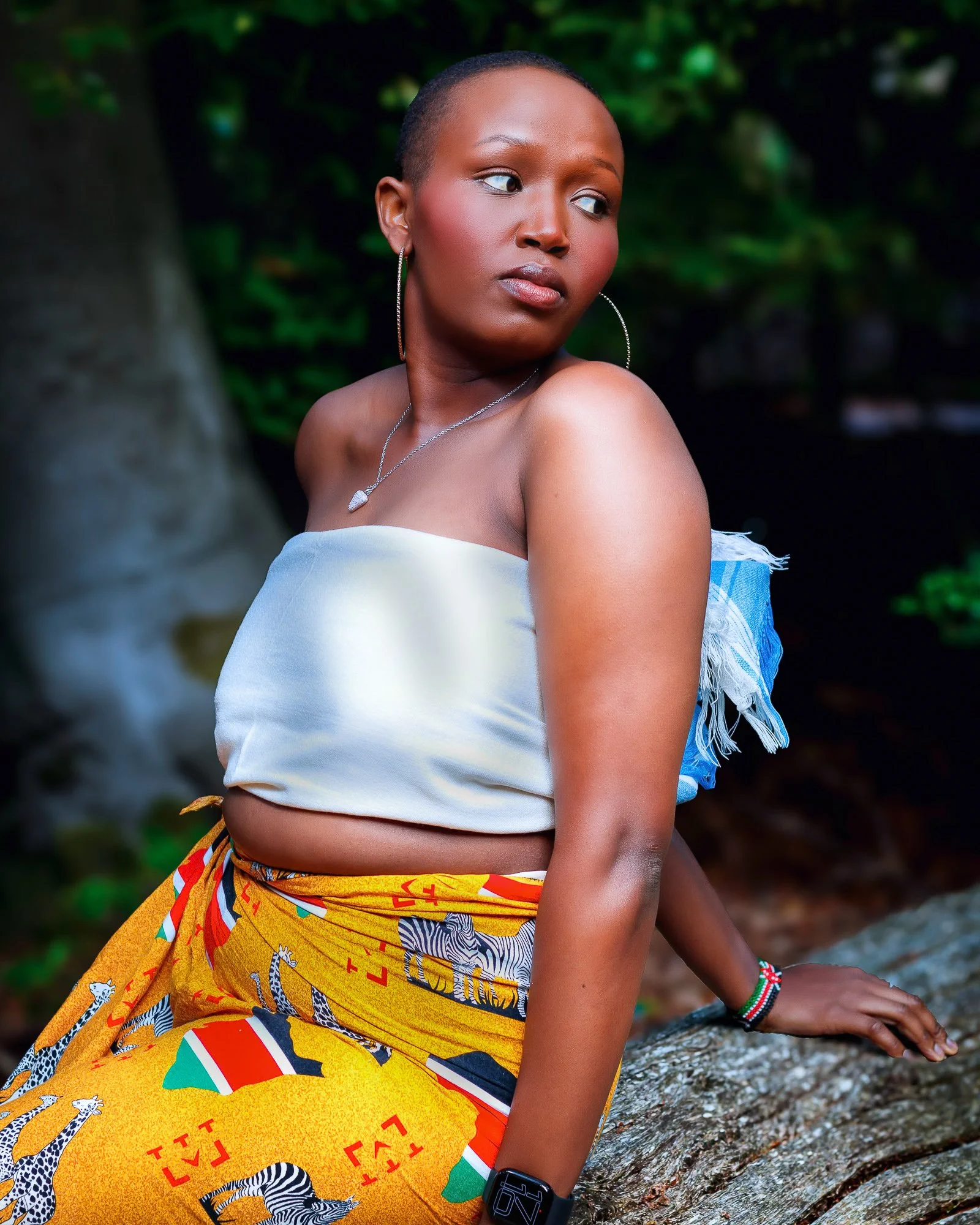 A woman with short hair and hoop earrings looking to her right, wearing a strapless white top, colorful patterned skirt, and accessories, outdoors with greenery in the background.