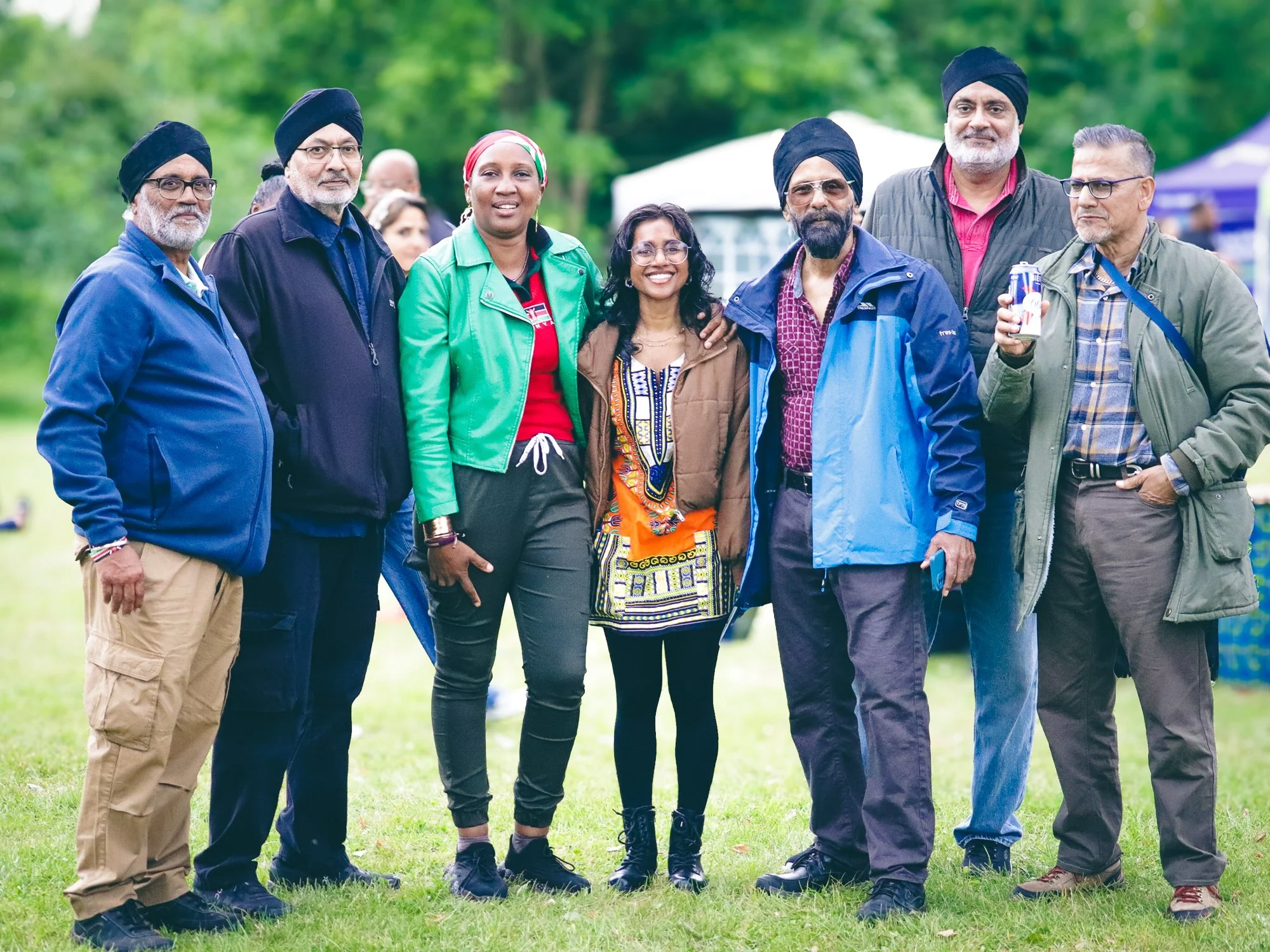 Group of seven diverse adults standing outdoors on a grassy field, smiling, with tents and trees in the background.