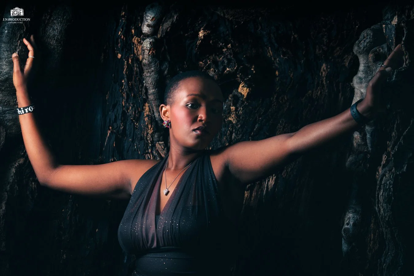A woman with dark skin, short hair, dressed in a black, glittery dress, posed against a dark, textured rock background, with her arms outstretched and looking confidently at the camera.