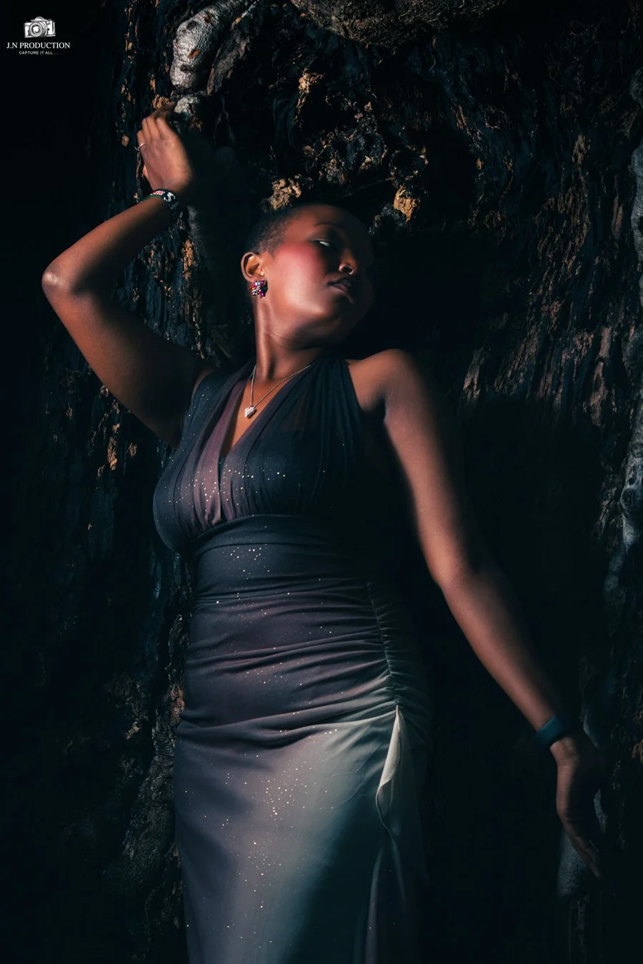 A woman in a dark, glittering gown leaning against a textured, rocky surface with her eyes closed and her arm raised above her head, wearing jewelry including earrings, a necklace, and a bracelet.