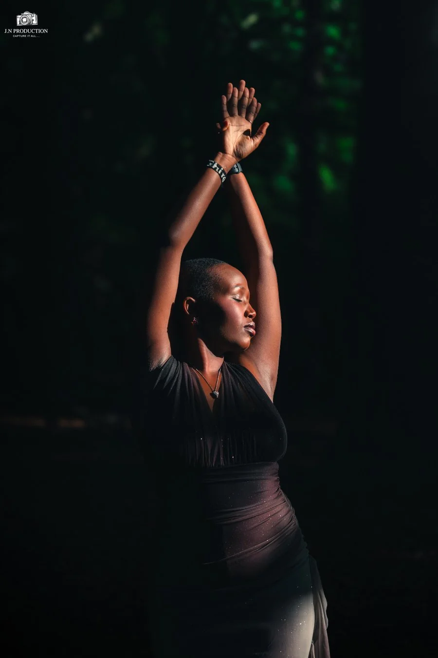 A woman with short hair dances or poses with her arms raised above her head, eyes closed, wearing a black dress and jewelry, against a dark background with faint green lighting.