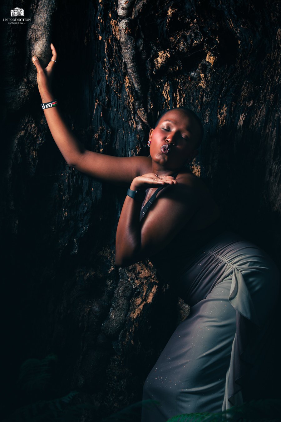 A woman with short hair making a kissy face pose, standing against a rocky, textured background, wearing a black top, flowy skirt, and jewelry, with her hand touching the rock wall.