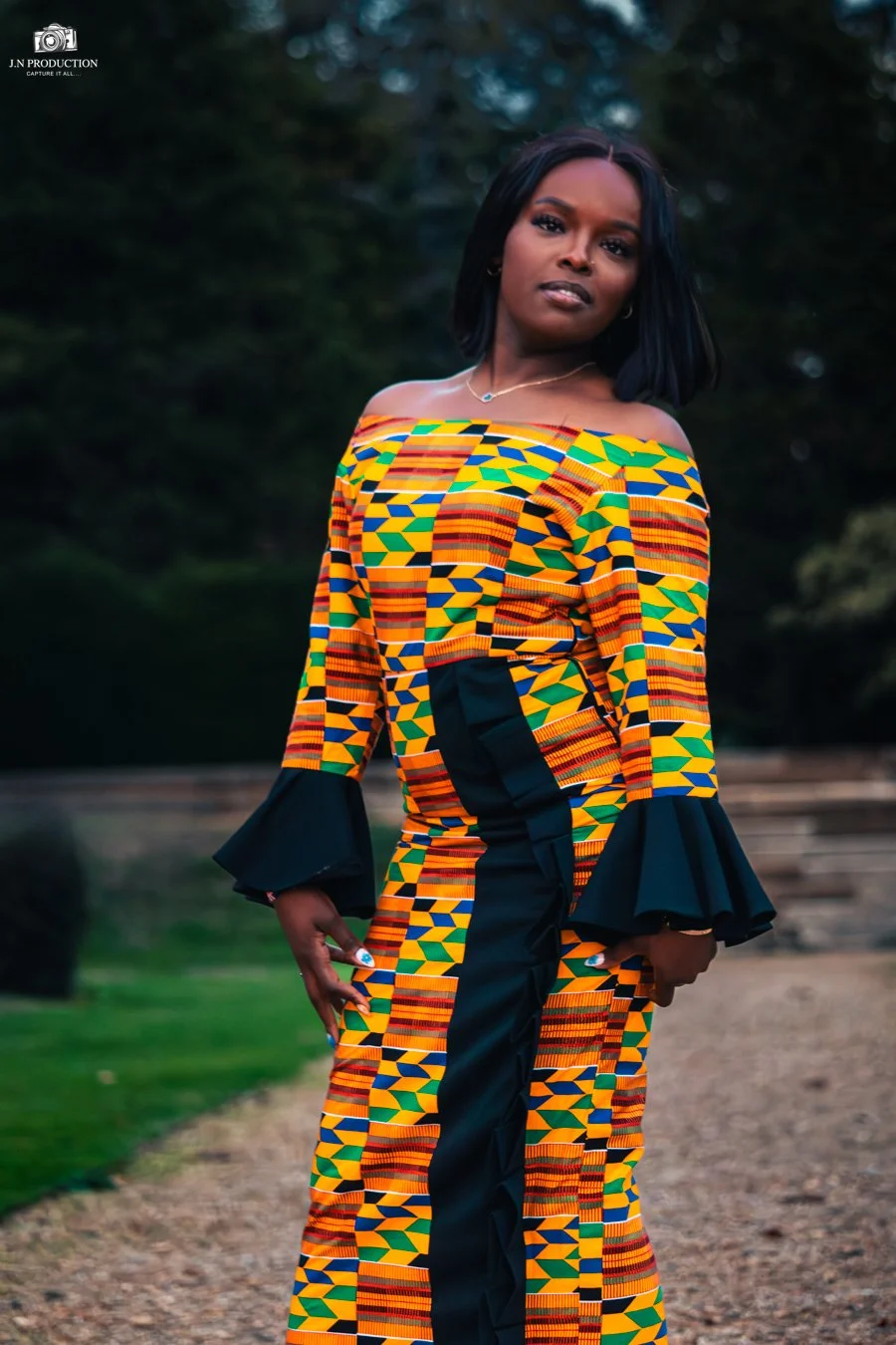 Woman in a vibrant, multicolored traditional African dress with geometric patterns, standing outdoors on a dirt path with trees in the background.
