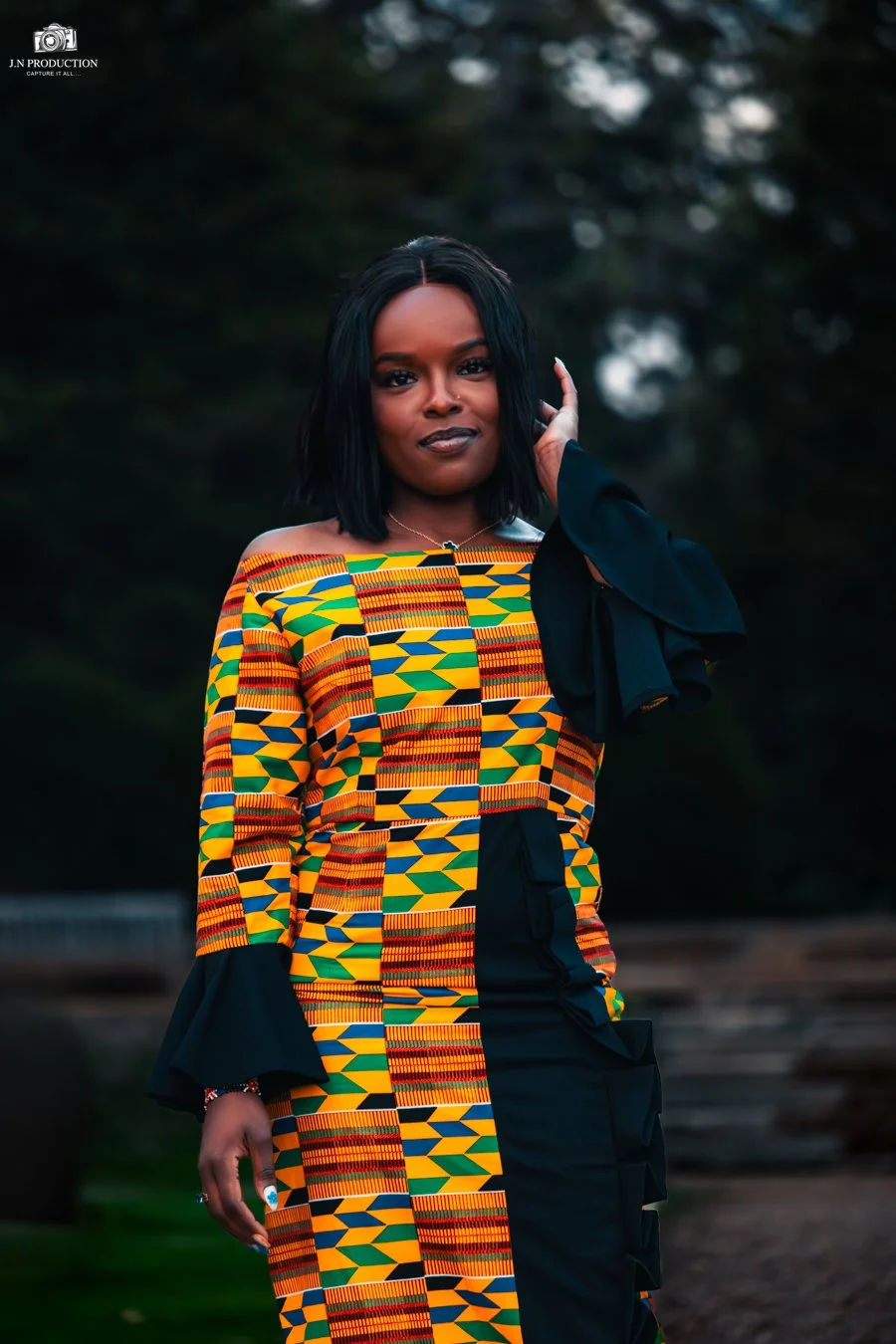 A woman with shoulder-length black hair, wearing traditional African print clothing with bright yellow, green, blue, and red geometric patterns, poses outdoors with a forested background.