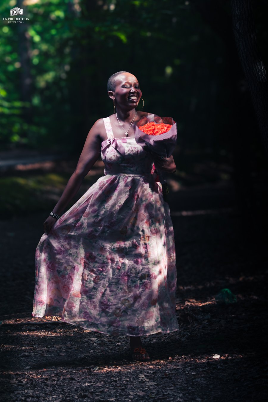A woman with short hair, wearing a sleeveless, floral dress, is holding a bouquet of orange flowers in a shaded outdoor area with trees and greenery in the background.