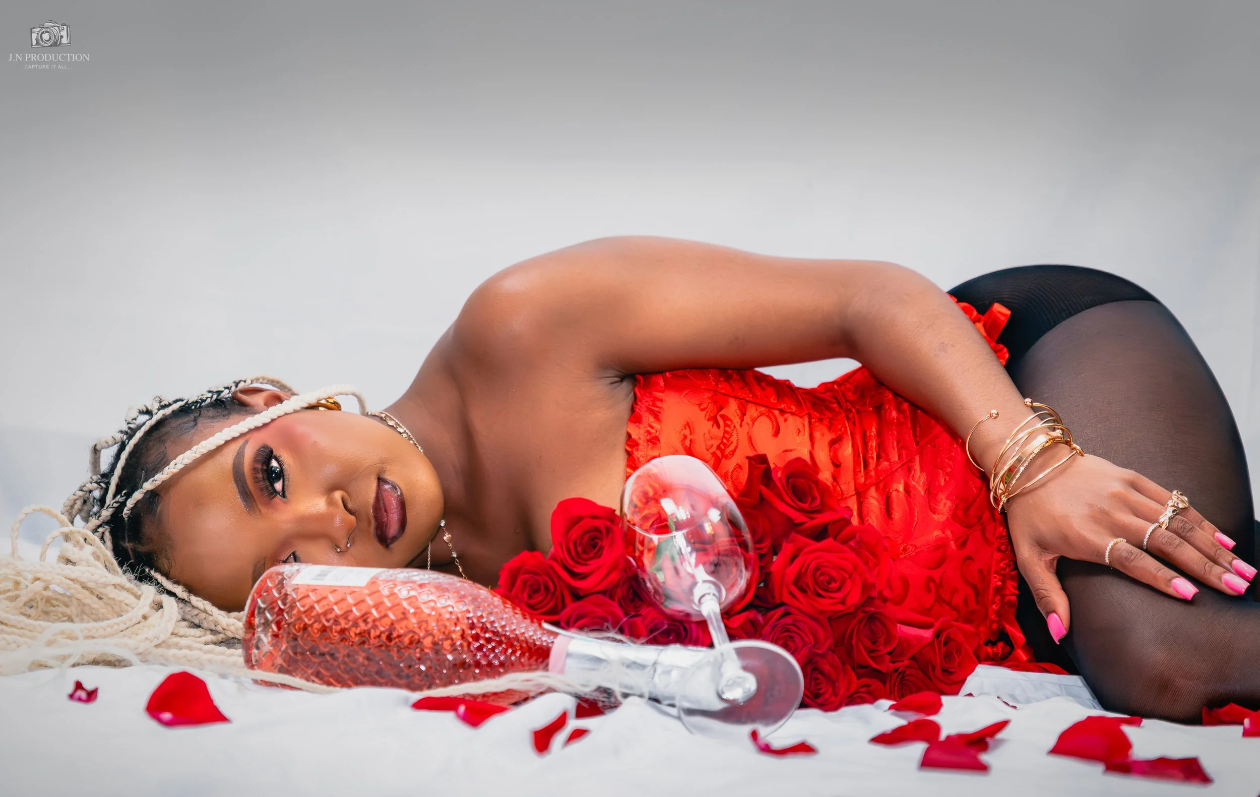 Woman with braided hair, lying on her side on white sheet, surrounded by red roses, wine glass, and decorative glass bottle, wearing red dress and jewelry.