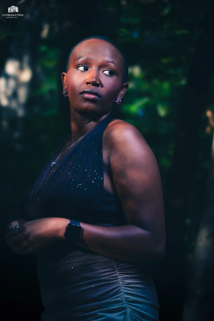 A woman with short hair dressed in a sparkly dark blue top, looking to her left, outdoors with greenery in the background.