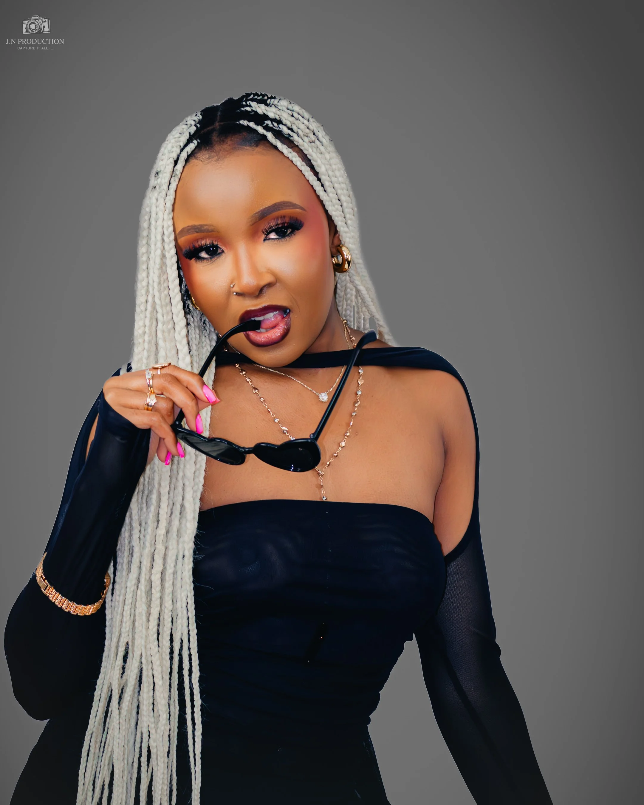 Woman with long, white braids wearing a black off-shoulder dress and holding sunglasses, posing against a plain gray background.