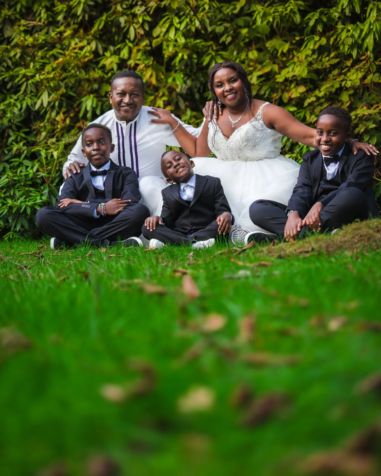 A family photo of six people sitting on the grass, with a lush green bush in the background. An adult man and woman are in the center, smiling. They are surrounded by three young boys in suits, also smiling and sitting cross-legged on the ground.