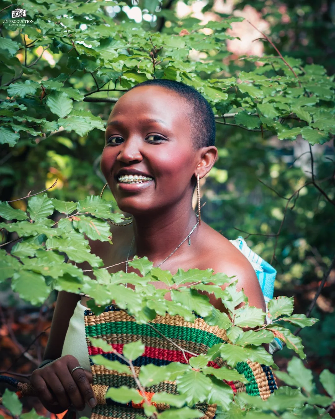 A young woman with short hair and earrings smiling through green leaves in an outdoor natural setting.