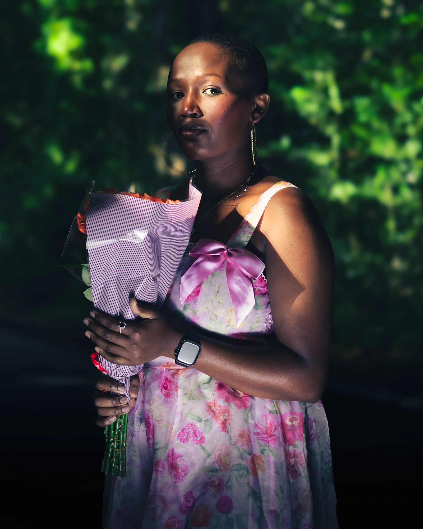 A woman with a short haircut holding a bouquet of flowers, wearing a floral dress with a pink bow, accessorized with earrings and a smartwatch, standing outdoors with a background of blurred green foliage.