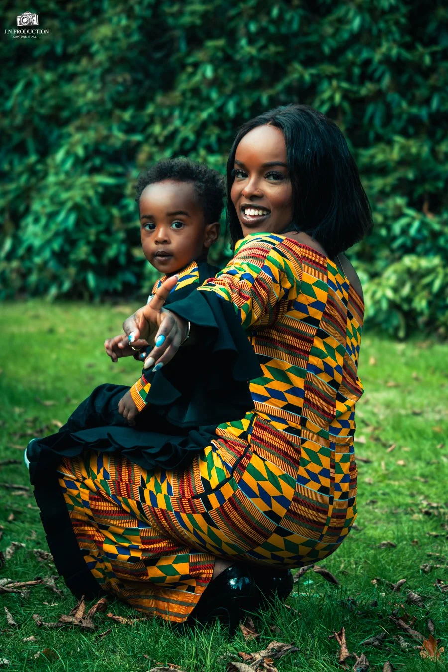 A woman and a young child sitting outdoors on grass, both wearing vibrant, colorful kente cloth clothing, with lush green foliage in the background. The woman is smiling and pointing at the camera, while the child looks at the camera with a neutral e