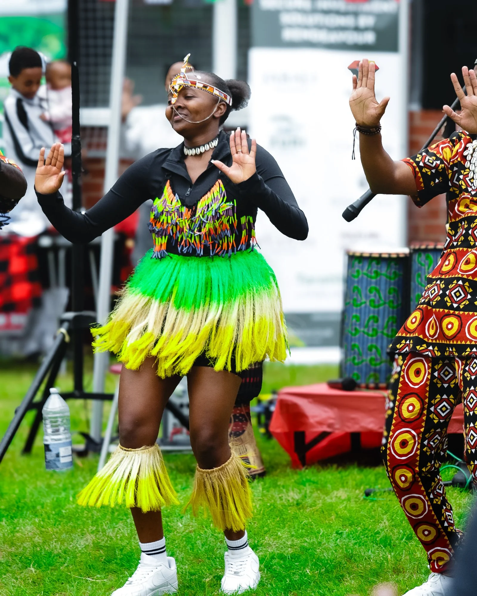 A woman in traditional African attire dancing outdoors during a cultural event, wearing a colorful dress made of beads and feathers, with matching ankle and wrist adornments.