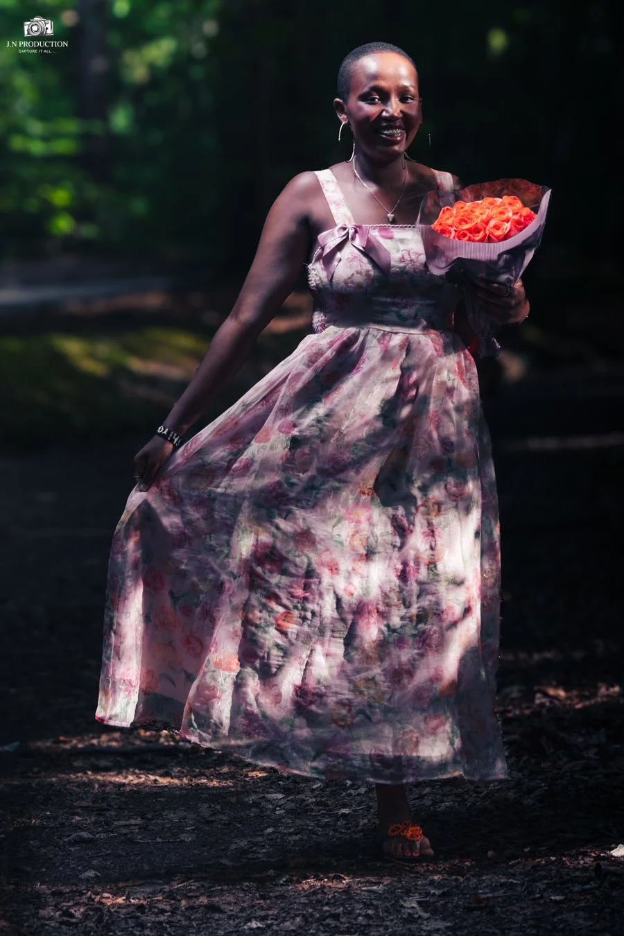 Woman in a floral dress holding a bouquet of orange roses, walking outdoors in a wooded area.