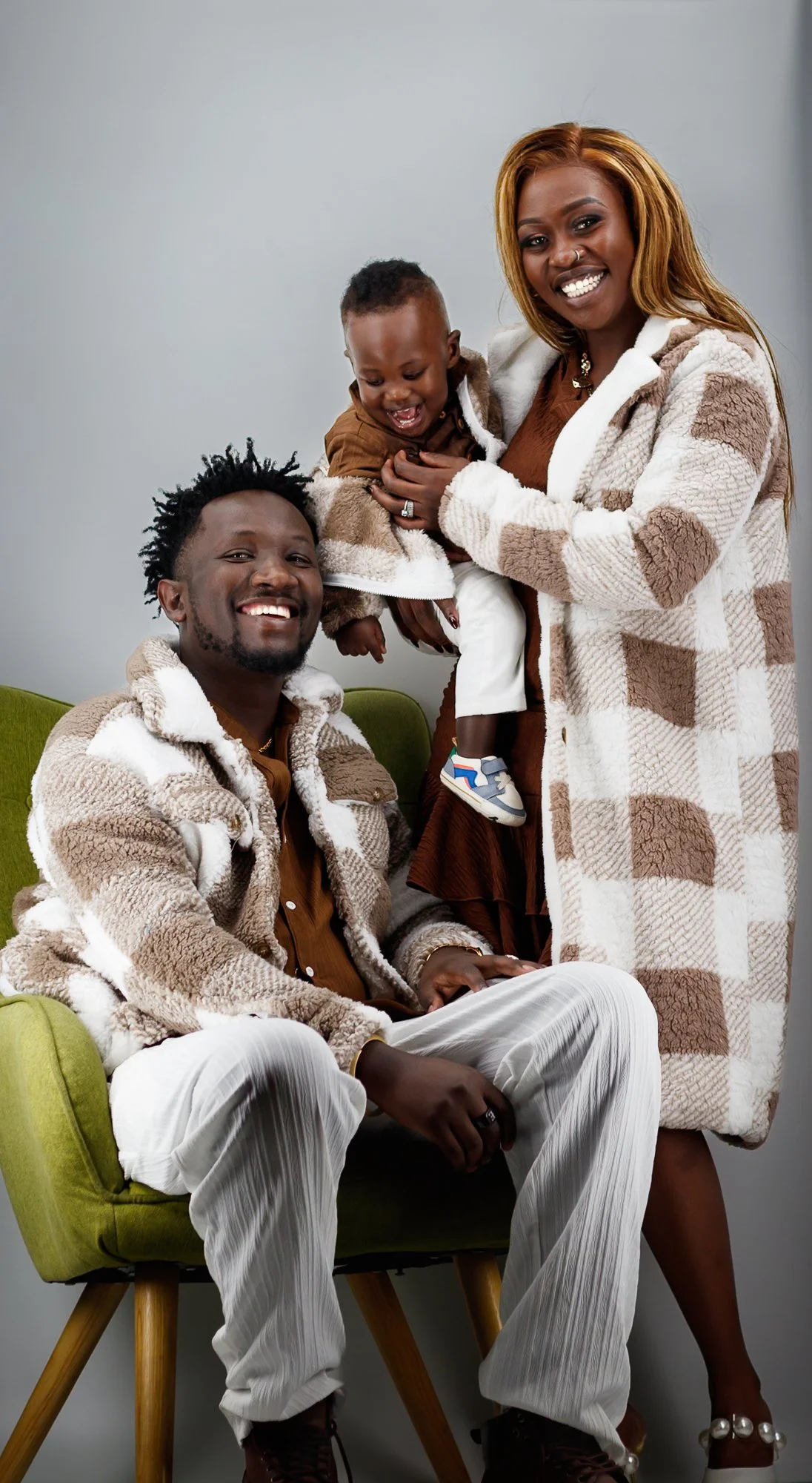 A joyful family of three together indoors. The woman is holding the child while standing, and the man is seated on a green chair, all smiling and wearing cozy, patterned jackets.