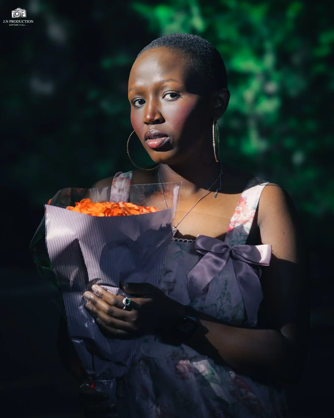 A woman with short hair and hoop earrings holds a bouquet of orange roses, standing outdoors with a blurred green background.