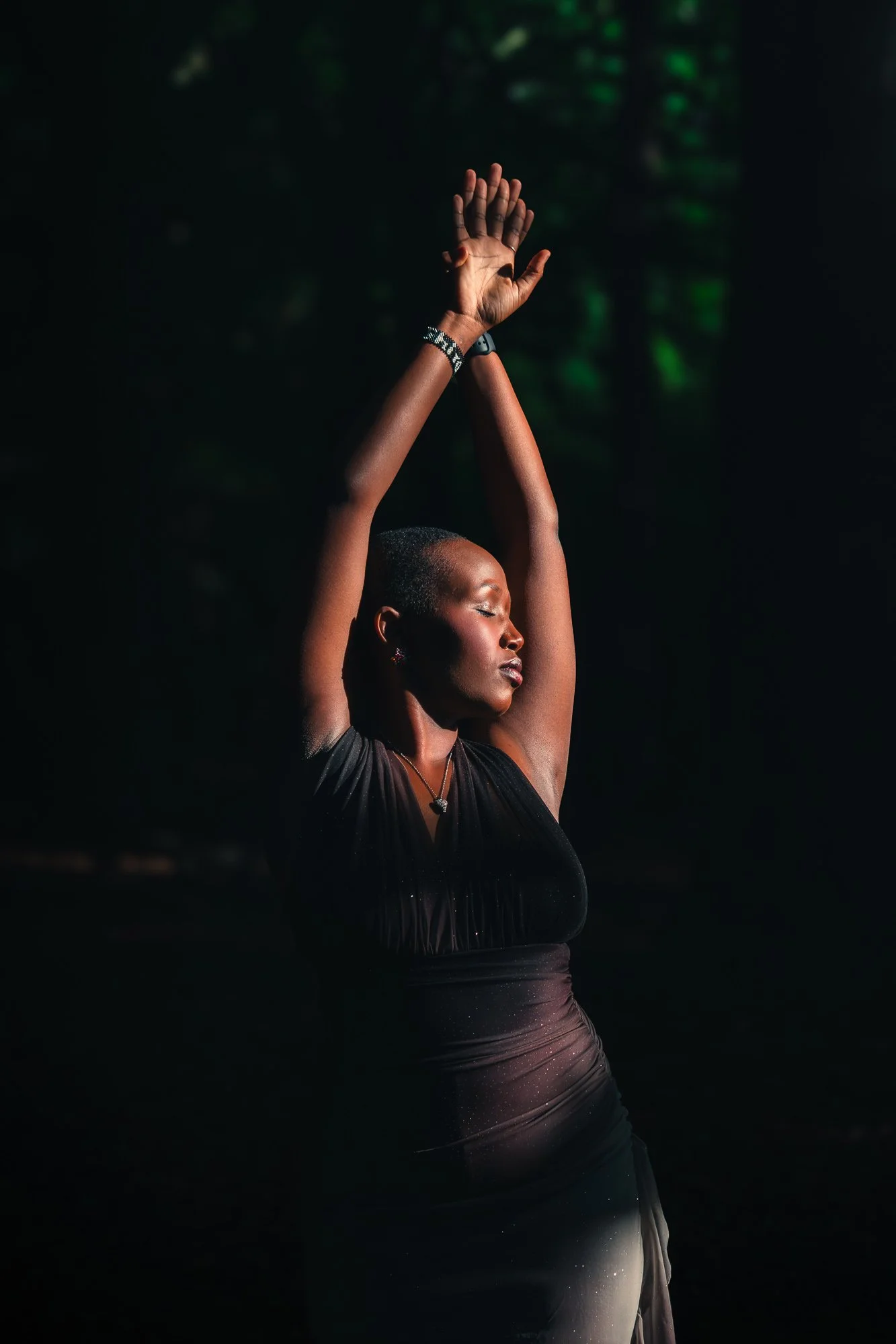 A woman with short hair and dark skin, wearing a black dress and jewelry, is standing with her eyes closed and arms raised above her head in a pose of serenity or dance, with a dark background and subtle green light.