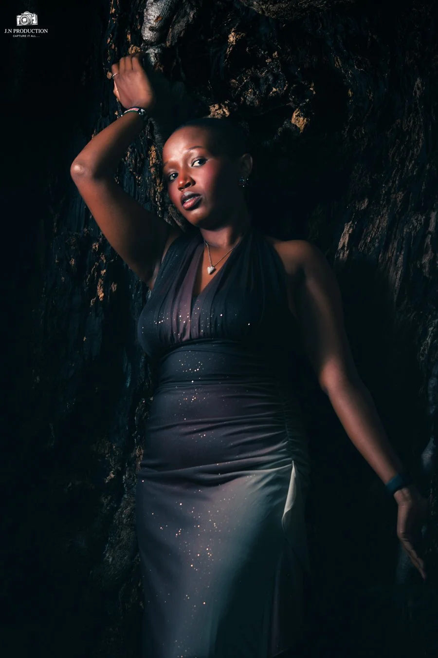A woman in a dark, elegant dress with sparkles, posing against a rocky, textured background with her arm raised above her head and her gaze directed at the camera.