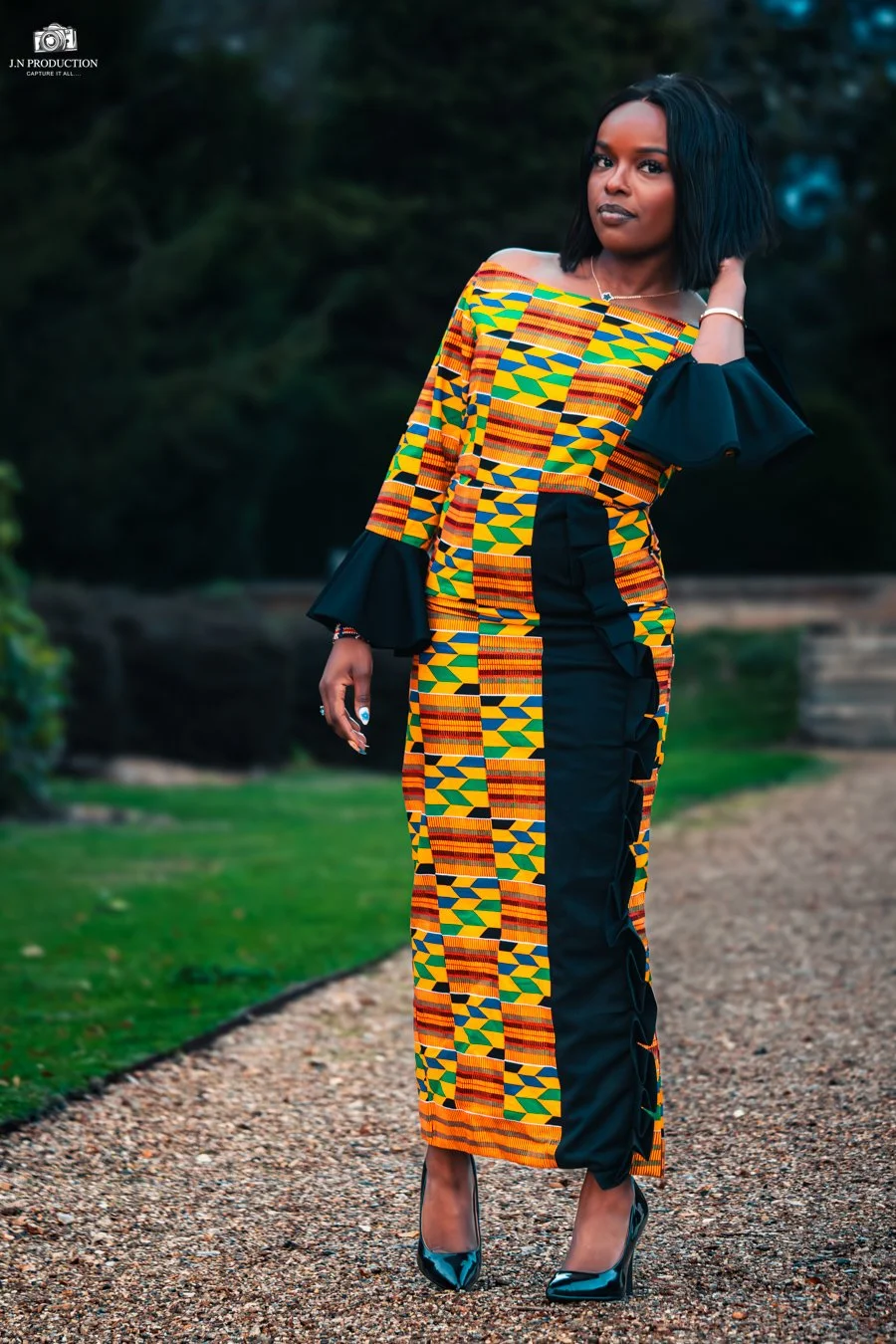 A woman in a colorful African print dress with black sleeves and high heels standing outdoors on a gravel path with greenery in the background.