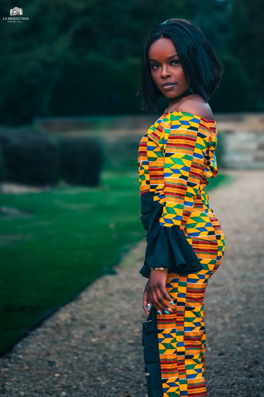A woman with shoulder-length black hair wearing a colorful, patterned dress with yellow, green, red, and blue geometric shapes, standing outdoors on a gravel pathway with greenery in the background.