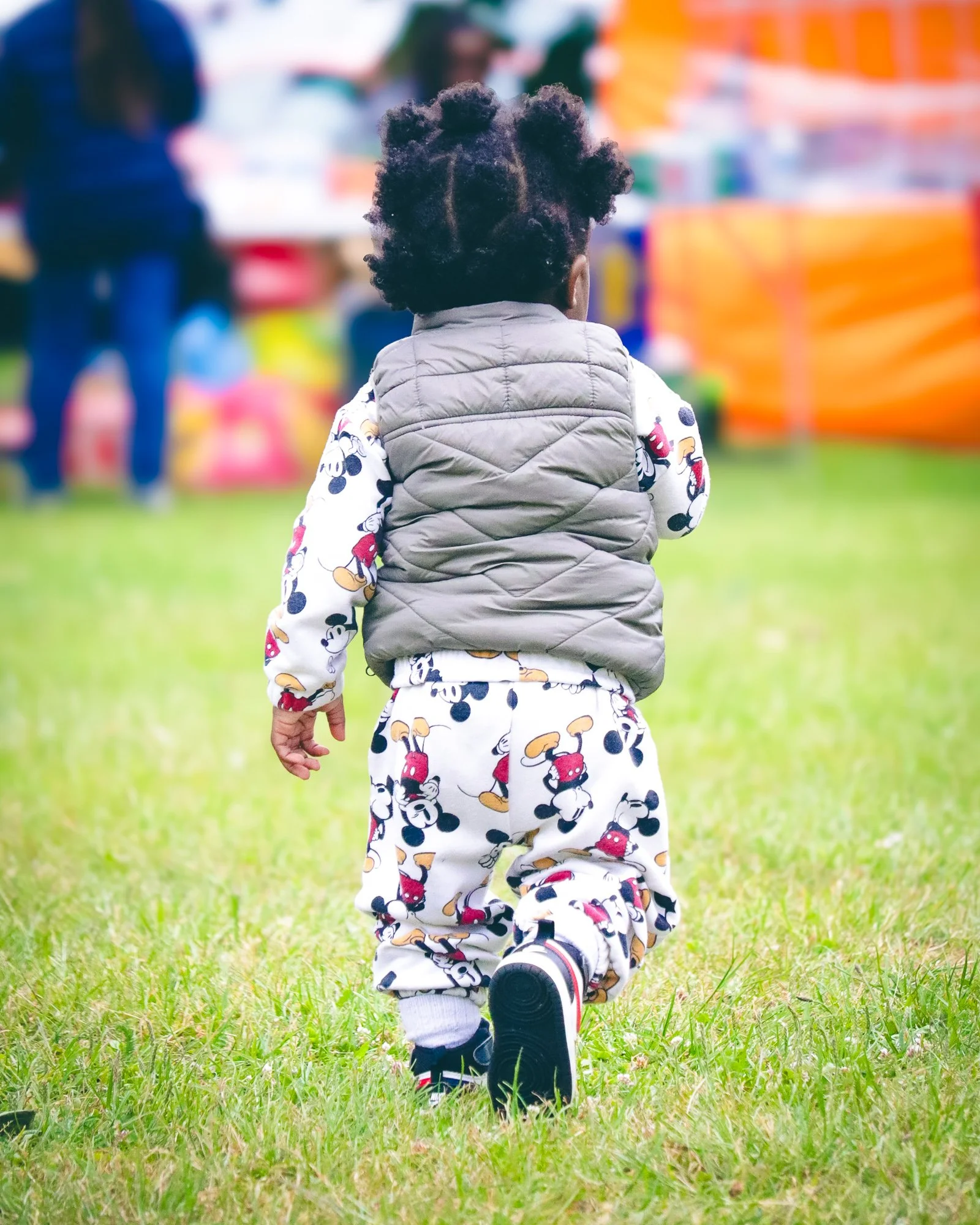 A young child with curly hair wearing a Mickey Mouse outfit and a grey vest, walking on grass at an outdoor event.