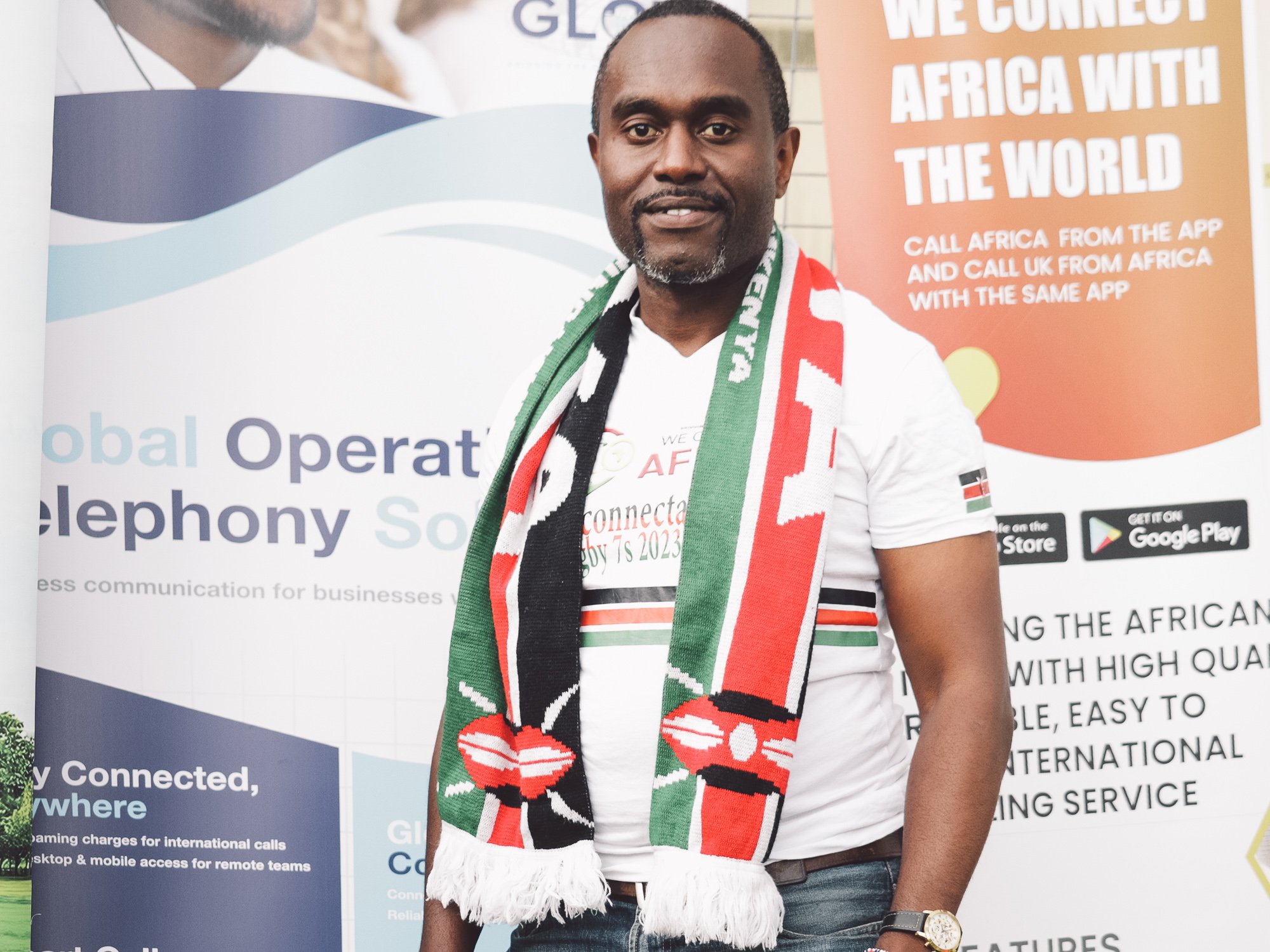 A man standing in front of a promotional banner, wearing a white t-shirt with black, red, and green details, and a colorful scarf with red, black, green, and white. He has a short beard, is smiling, and is wearing a watch on his left wrist.