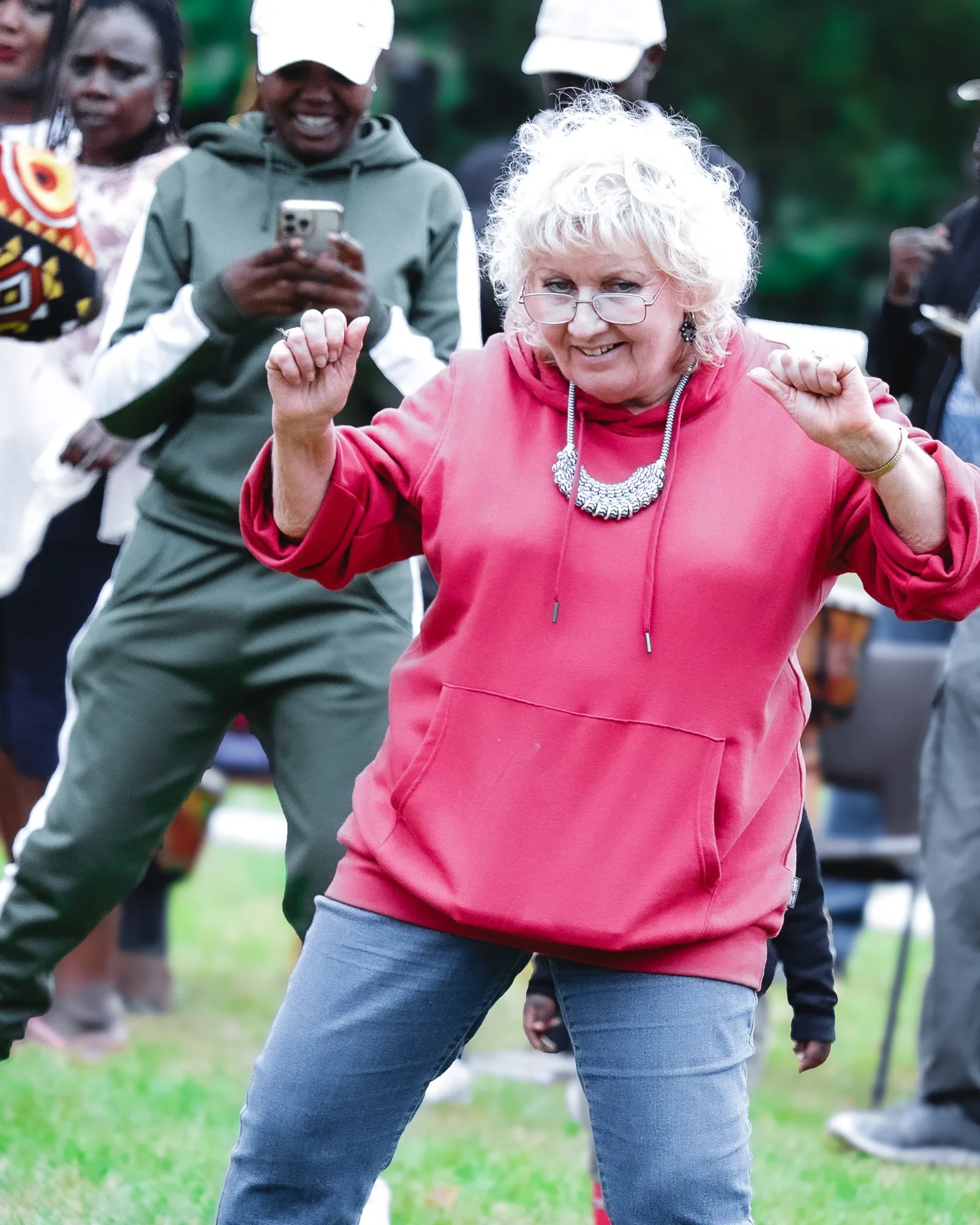 An elderly woman with white curly hair, glasses, and a pink hoodie dancing and smiling outdoors, with other people in the background.