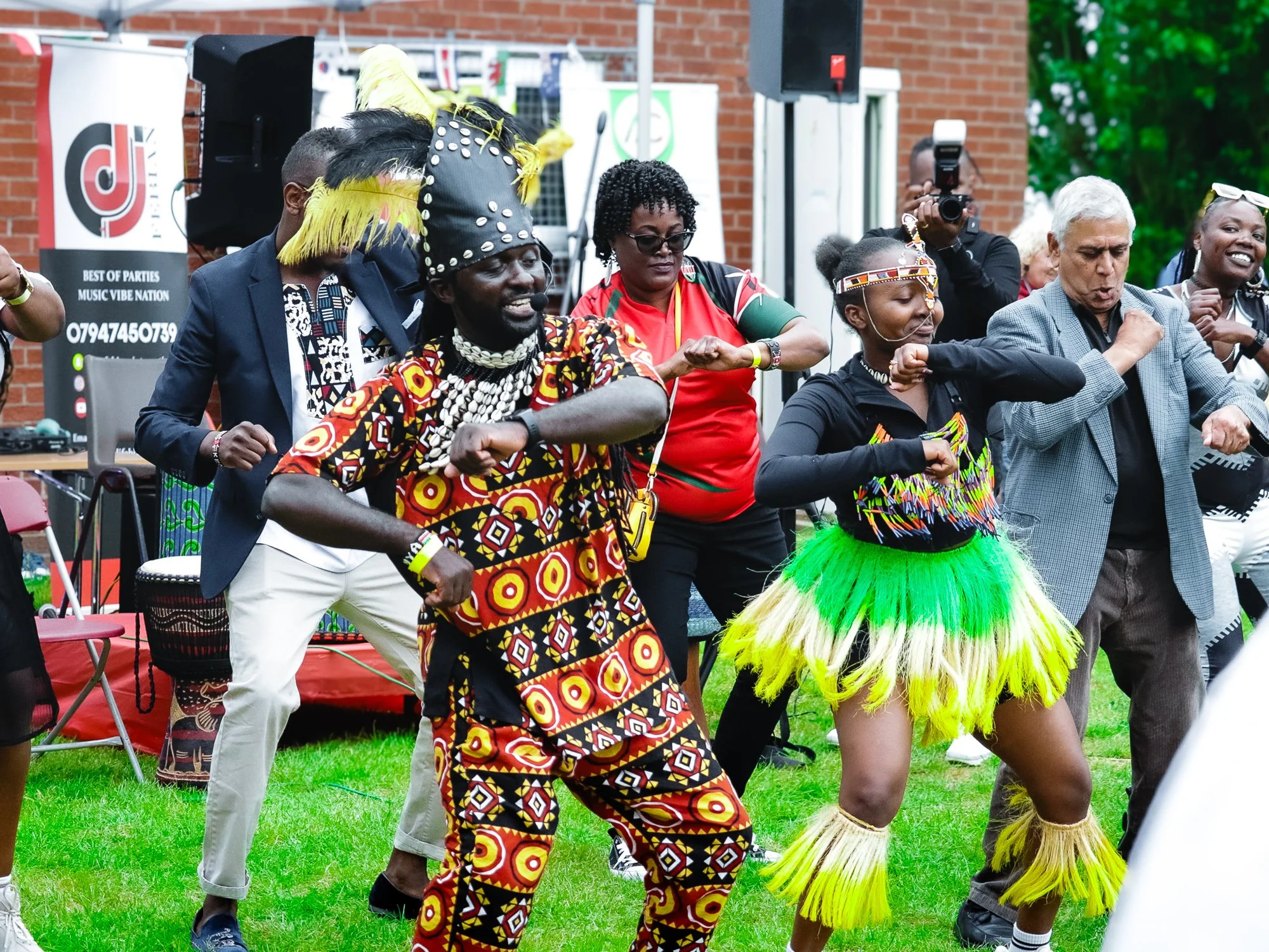 People dancing outdoors at a cultural event, dressed in traditional African attire, with some wearing colorful skirts, beads, and headpieces, while a DJ booth and photographer are visible in the background.