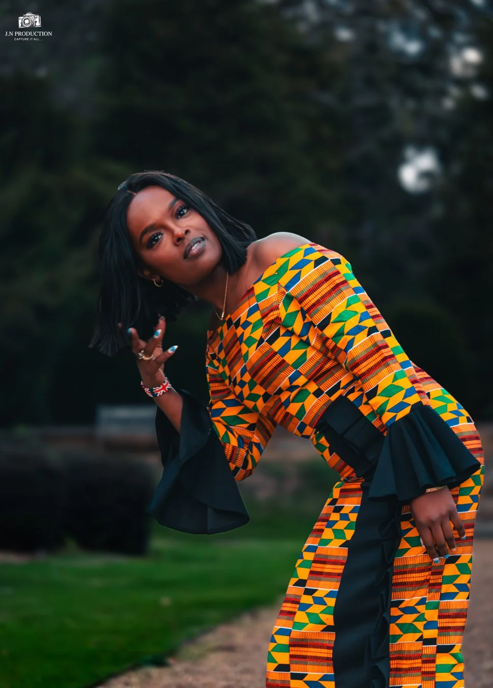 A woman in colorful African print clothing poses outdoors during daytime, with trees in the background.