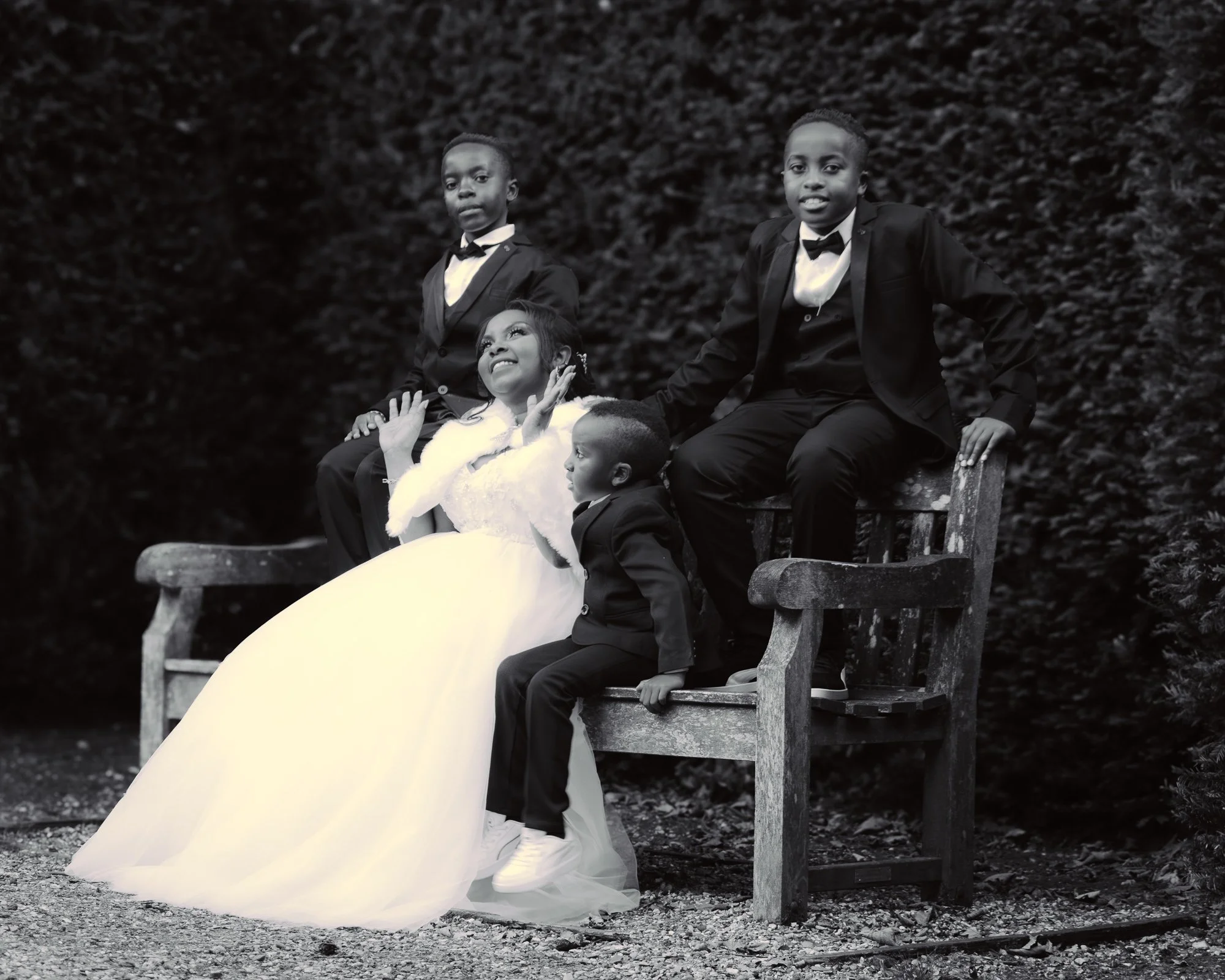 A black and white photo of five children dressed in formal attire, with two boys in tuxedos and three girls, one of whom is wearing a white wedding gown, posing outdoors on a wooden bench and tree stump surrounded by bushy trees.