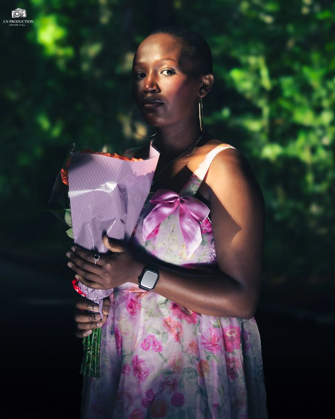 Woman with short hair wearing a floral dress, holding a bouquet of flowers, standing outdoors with greenery in the background.