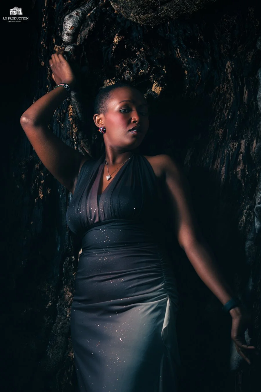 A woman wearing a black dress with glitter details, jewelry, and makeup, posing against a dark, rocky background with one arm raised and head tilted.
