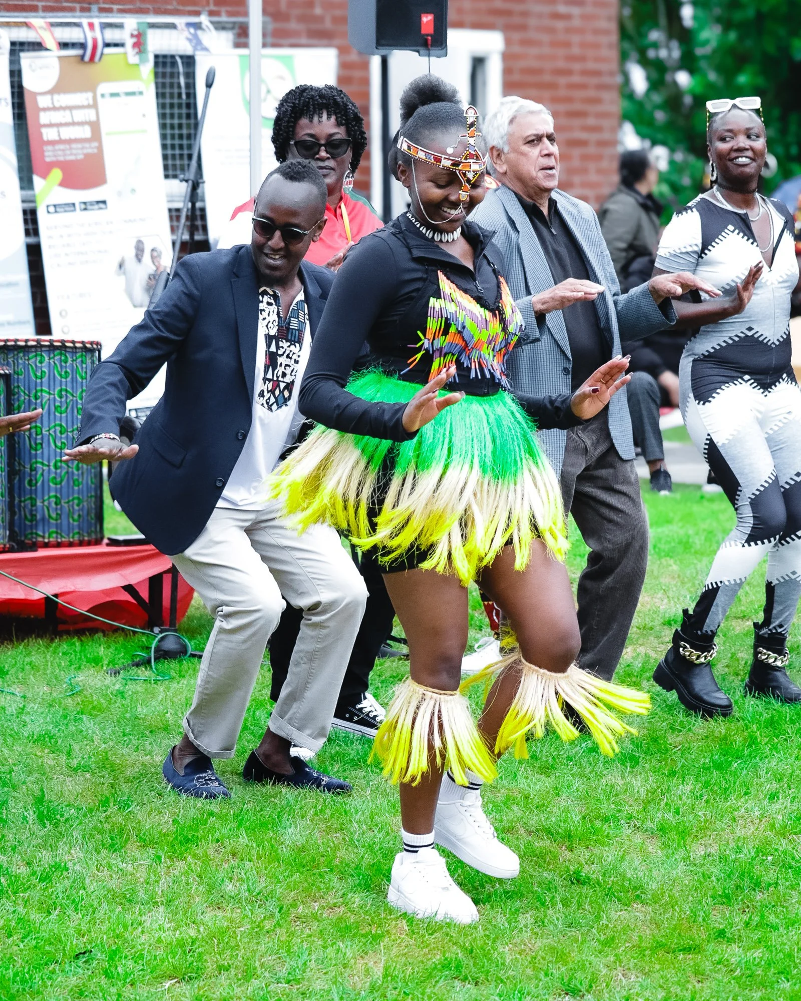 Group of people dancing outdoors, with one woman wearing a colorful, traditional outfit with grass and bead details, and others dressed in modern clothing.