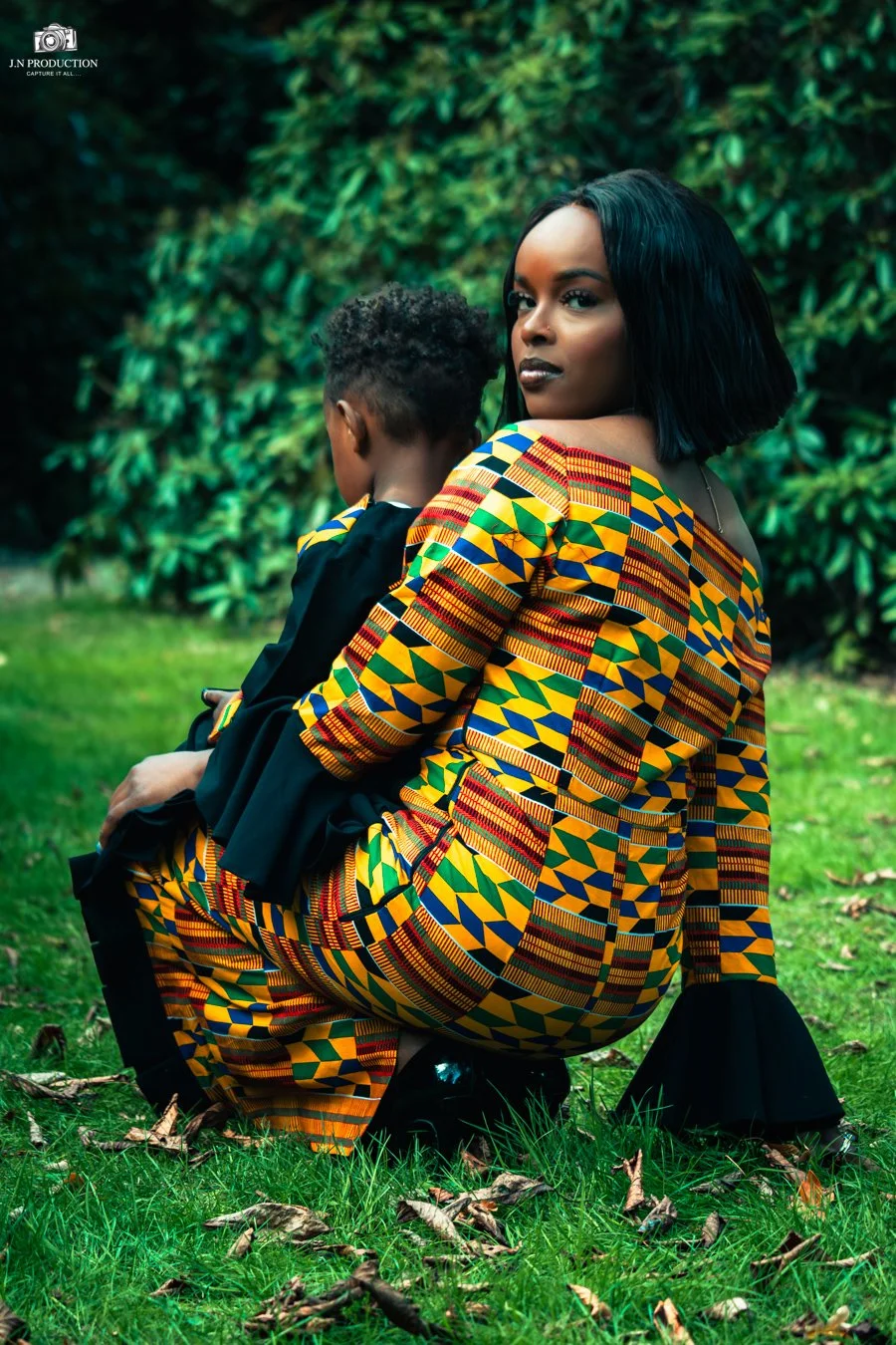 A woman in a colorful traditional African dress holds a young boy in a black outfit while sitting on the grass in a park with green foliage in the background.