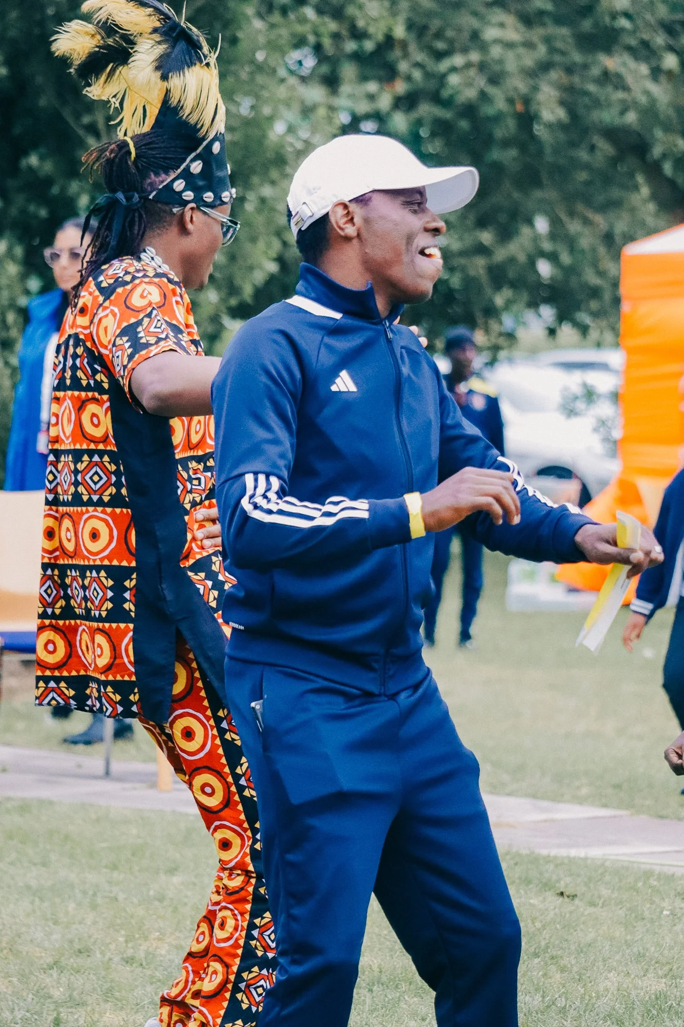 Two men outdoors, one in colorful African print attire with a feathered headpiece, the other in a blue tracksuit and white cap, smiling and holding a paper, during a daytime event.