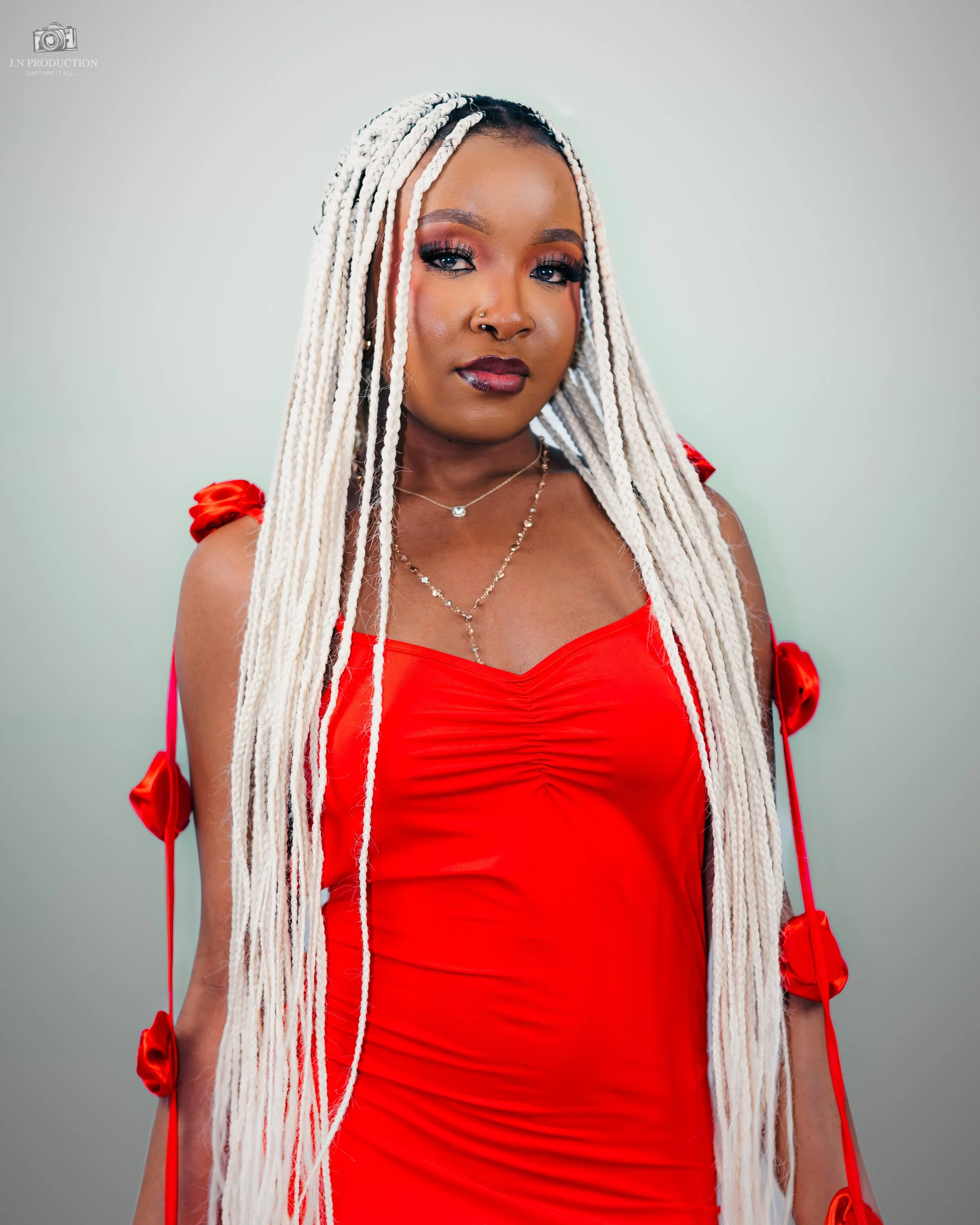 A woman with long white braided hair wearing a red dress with ribbon details, accessorized with layered necklaces, posing against a plain background.