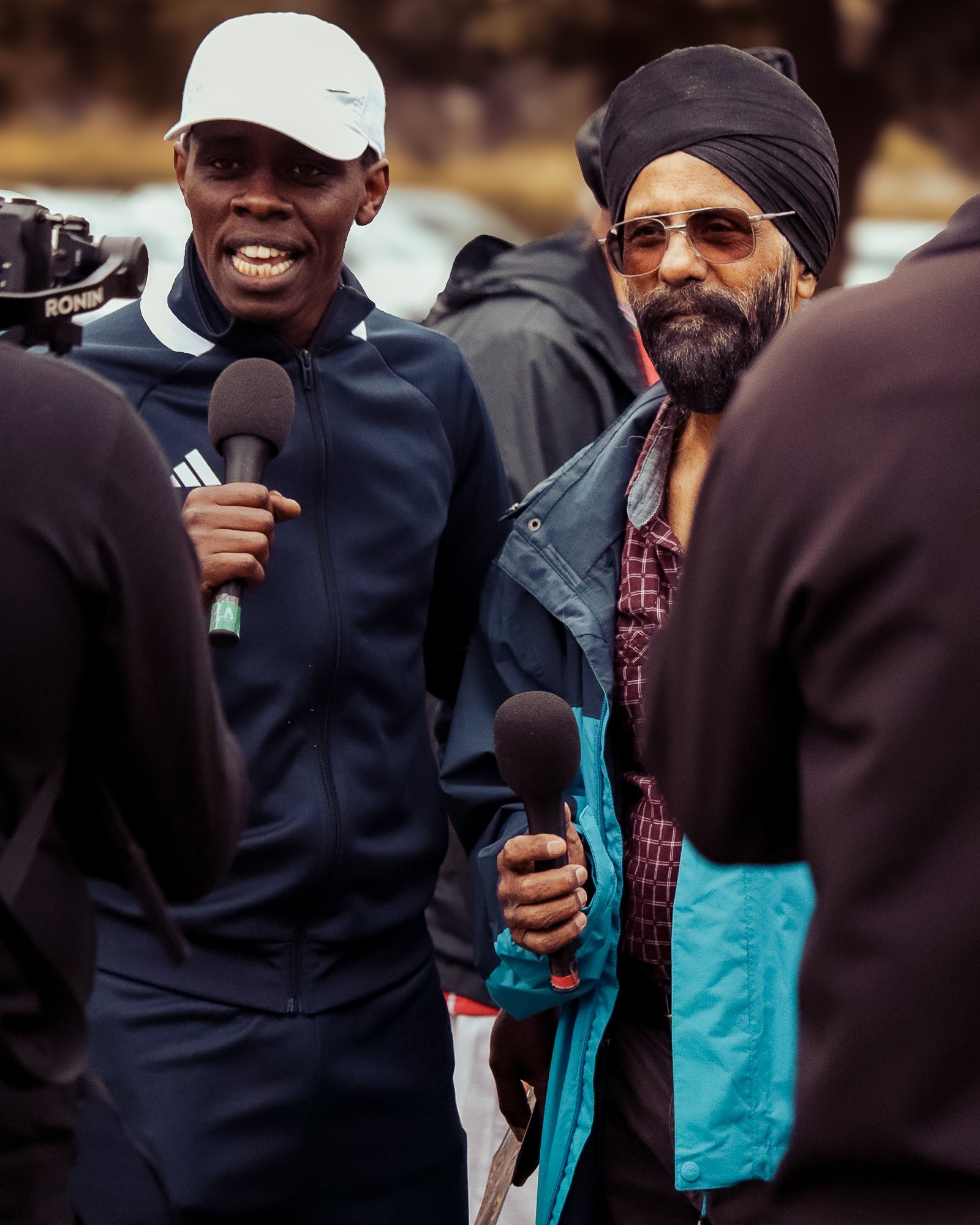 Two men holding microphones and speaking to reporters, one wearing a white cap and navy sporty jacket, the other wearing a black turban and sunglasses, outdoors during daytime.