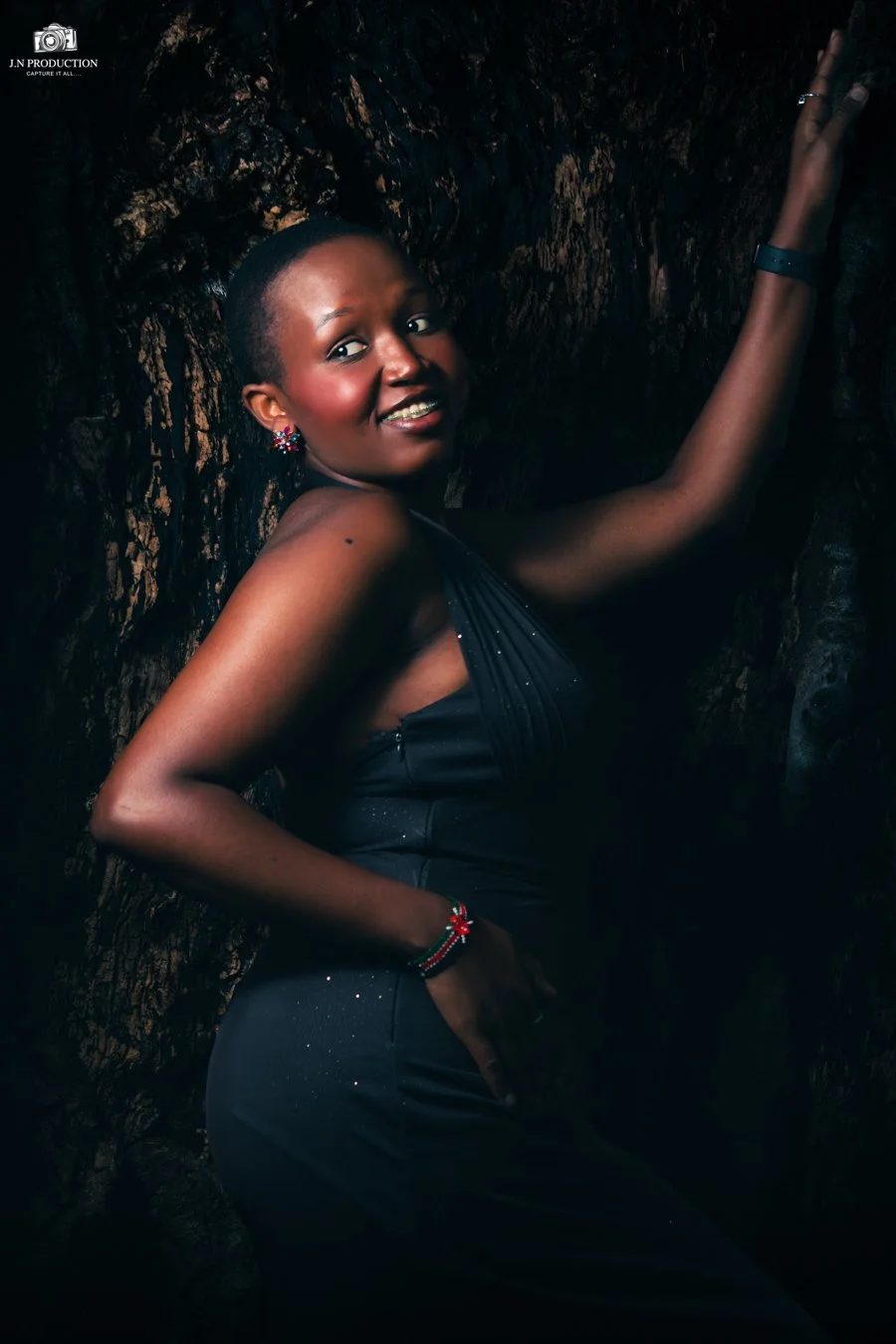 A smiling woman with short hair, wearing a black dress, standing against a dark, textured background, with one arm raised and the other resting on her hip.
