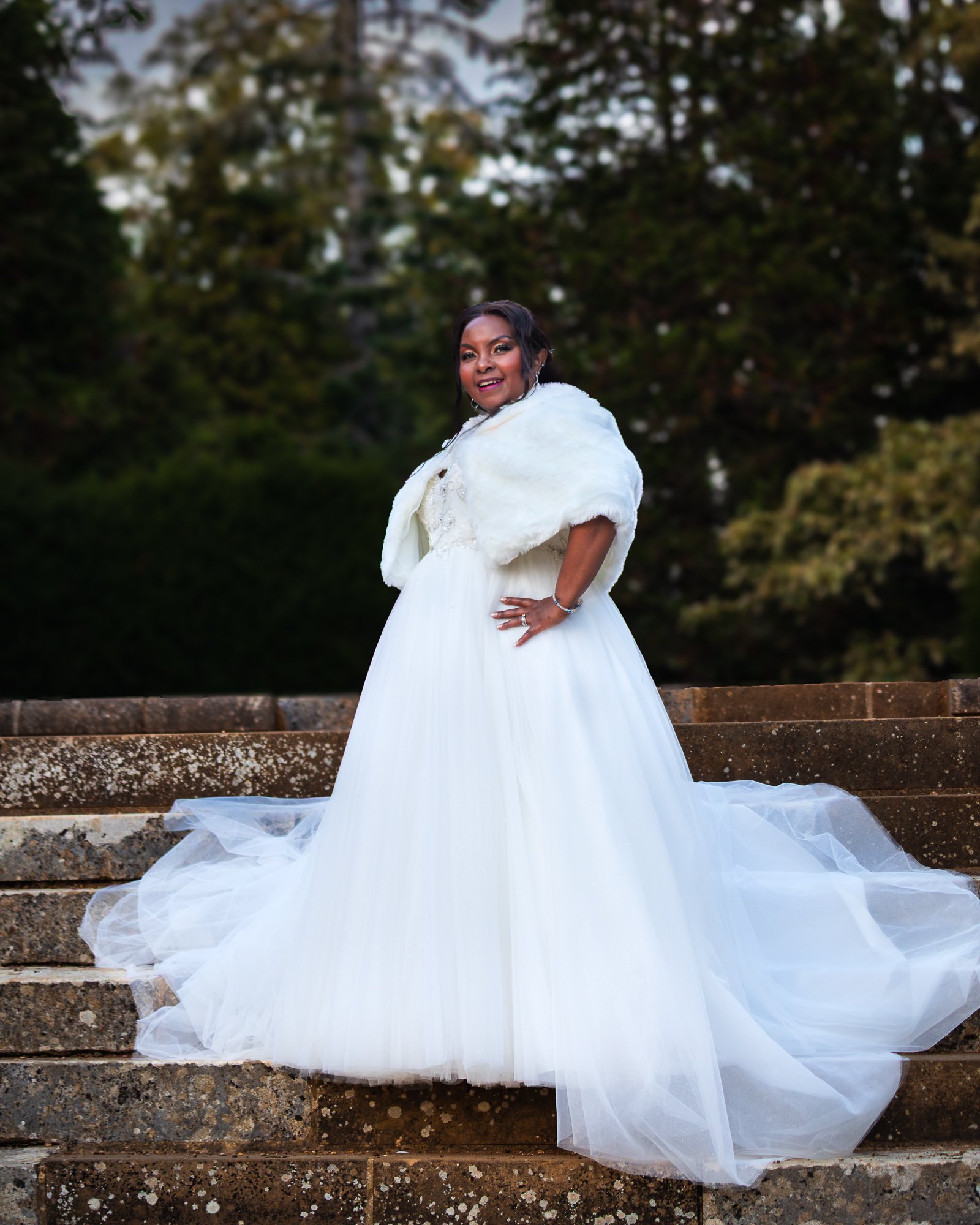 Woman in a white wedding dress and shawl standing on stone steps outdoors, smiling, with trees in the background.