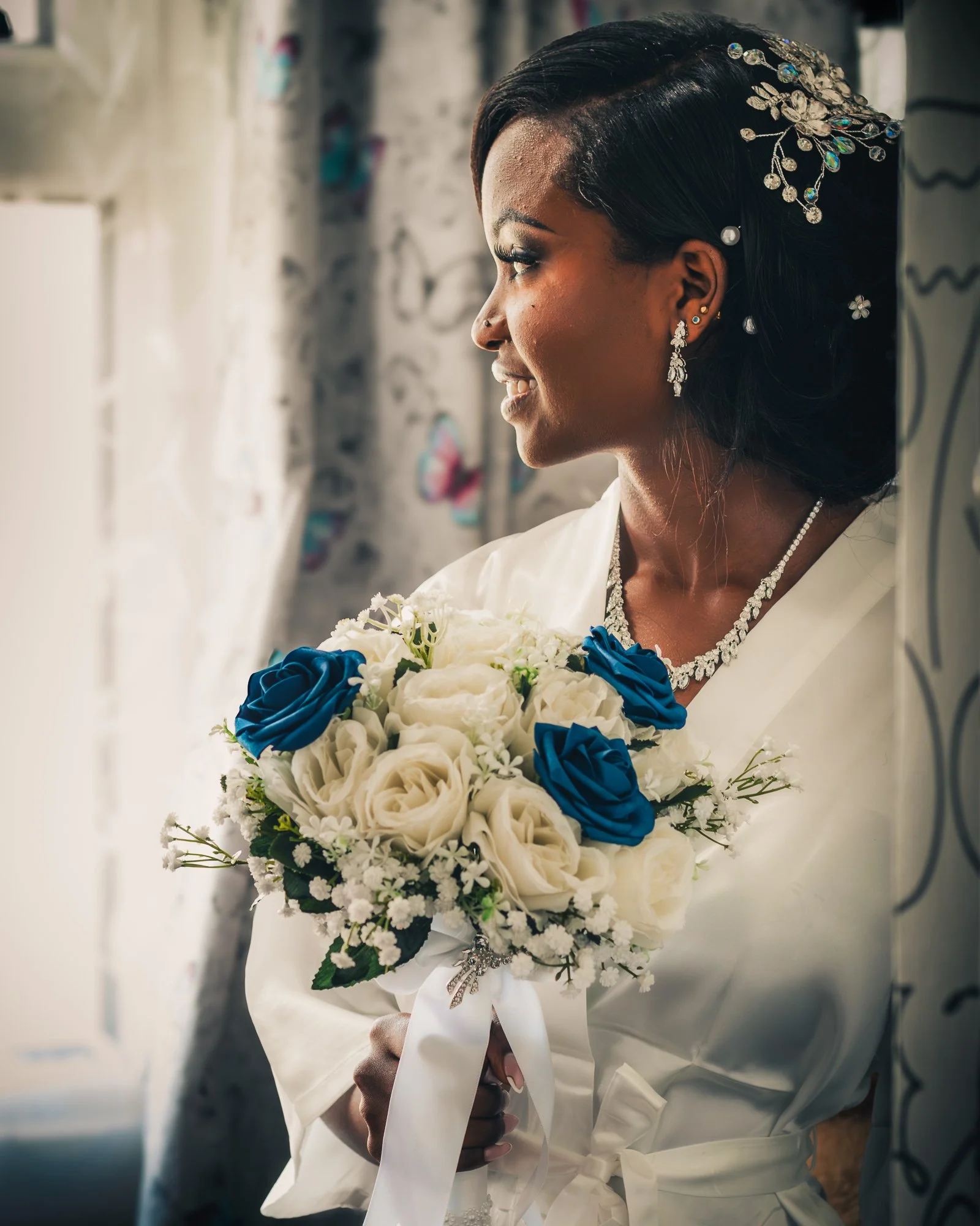 A woman in wedding attire holding a bouquet of white and blue roses, standing near a window with curtains.