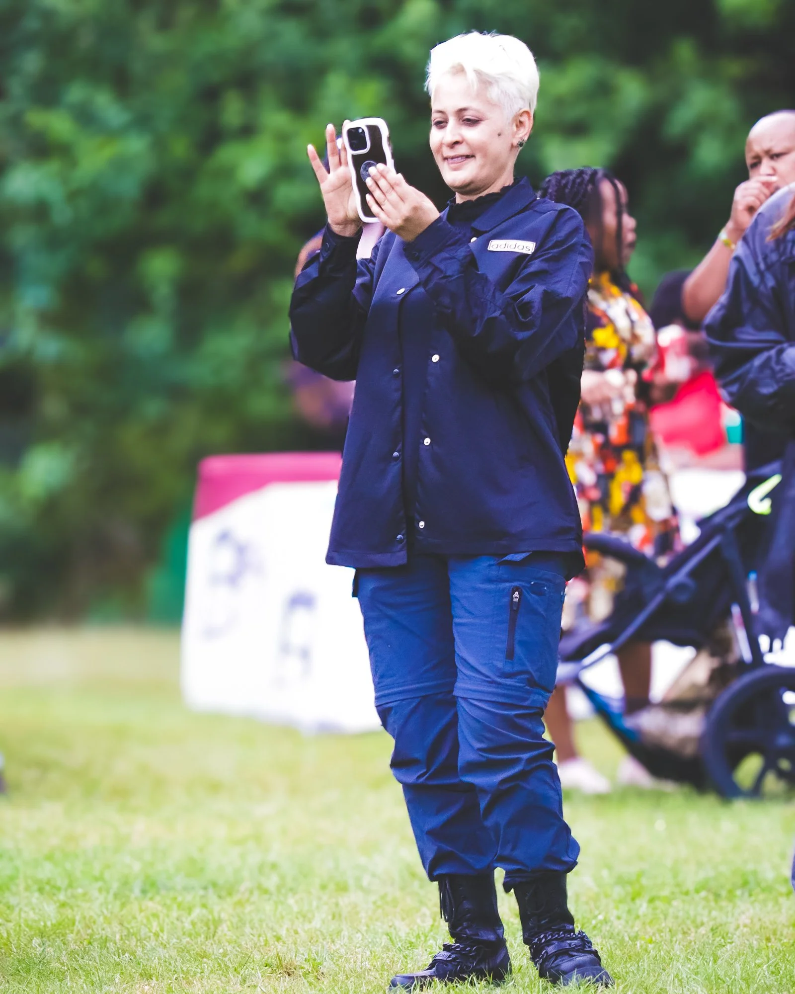 A person with short blonde hair wearing a dark blue jacket and cargo pants is taking a photo or video with a smartphone outdoors on a grassy field, with a group of people and trees in the background.