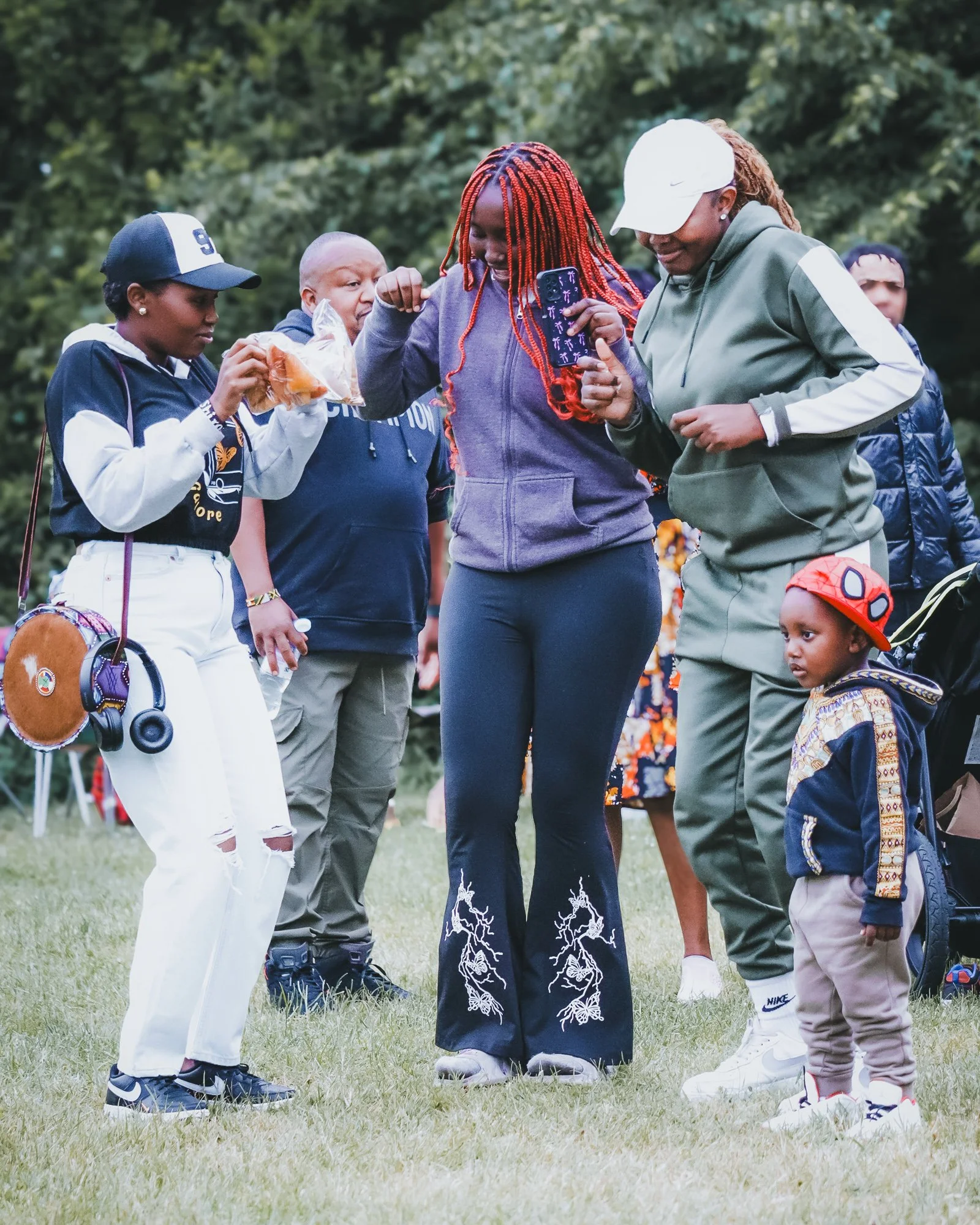 A group of five people standing outdoors on grass, with trees in the background. They are engaged in a conversation, with some holding snacks and taking pictures, including children and adults dressed casually.
