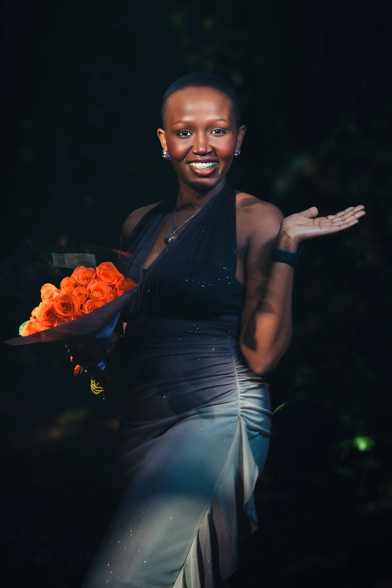 A woman smiling while holding a bouquet of orange roses, dressed in an elegant dark blue and gray evening gown, standing outdoors at night.