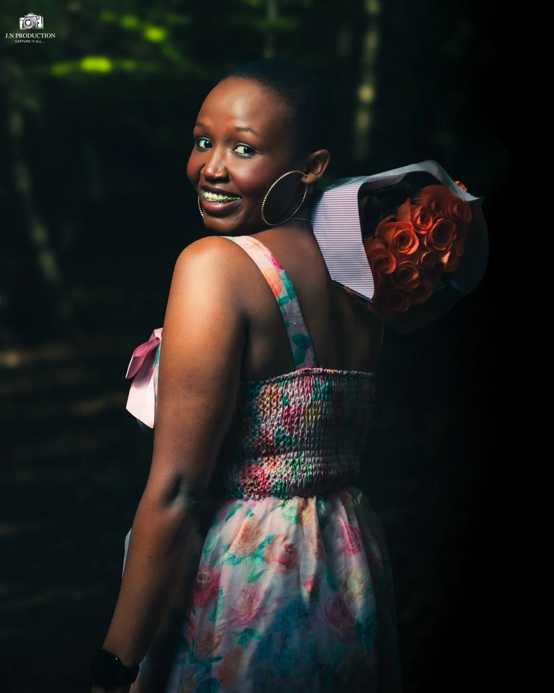 A woman smiling with braces, wearing a colorful floral dress and hoop earrings, holds a bouquet of orange roses over her shoulder in a forest setting.
