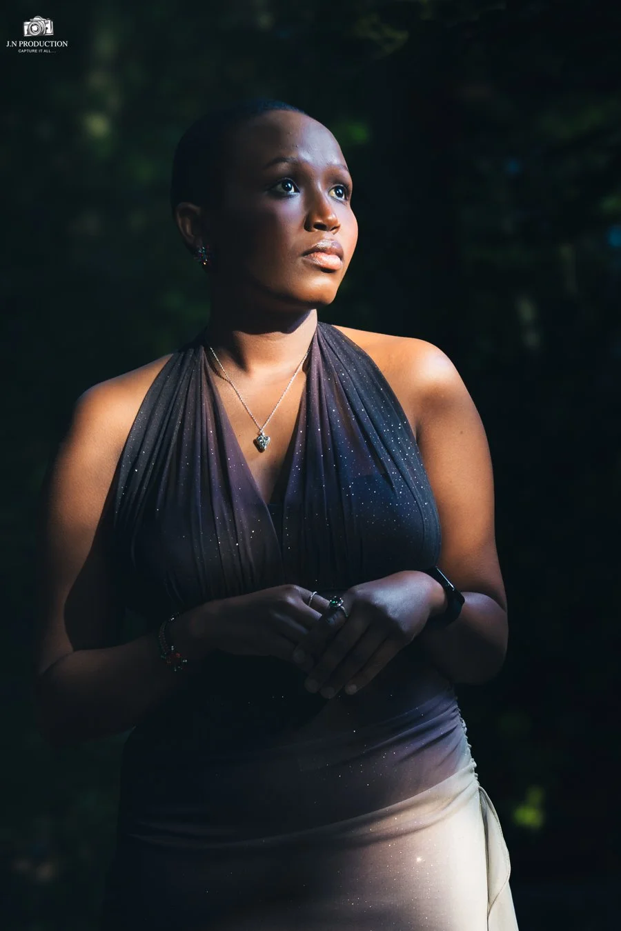 A woman with short dark hair and dark skin, wearing a sleeveless dark purple dress with a sparkly pattern, a silver necklace with a heart-shaped pendant, and multiple rings, stands outdoors in a natural setting with blurred greenery in the background