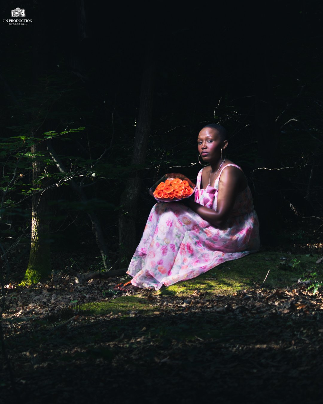 A woman in a pink floral dress sitting on the ground in a dark forest, holding a bouquet of orange roses.