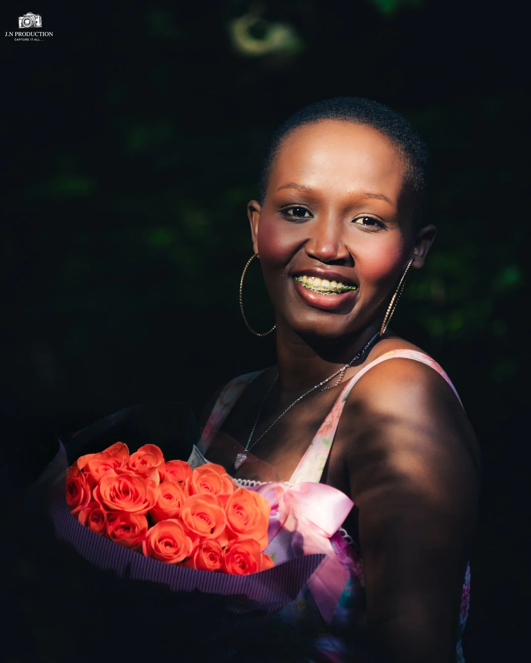 A smiling woman with short hair, wearing hoop earrings and a necklace, holding a bouquet of bright orange roses, standing outdoors with a blurred dark background.