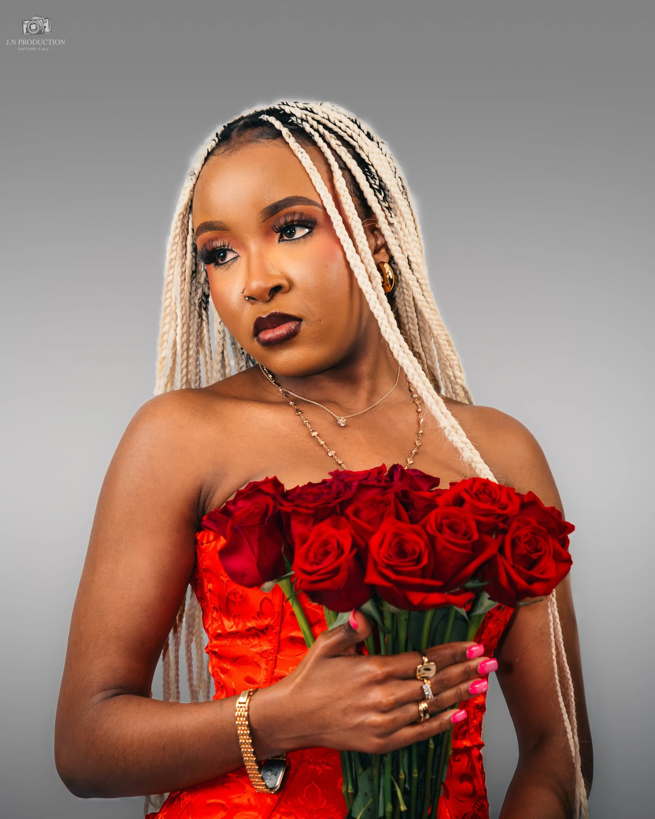 A woman with light blonde braids, wearing a red strapless dress, holding a bouquet of red roses. She has jewelry including necklaces, rings, earrings, and a bracelet, and is wearing makeup with dark lipstick and eye makeup. The background is plain gr