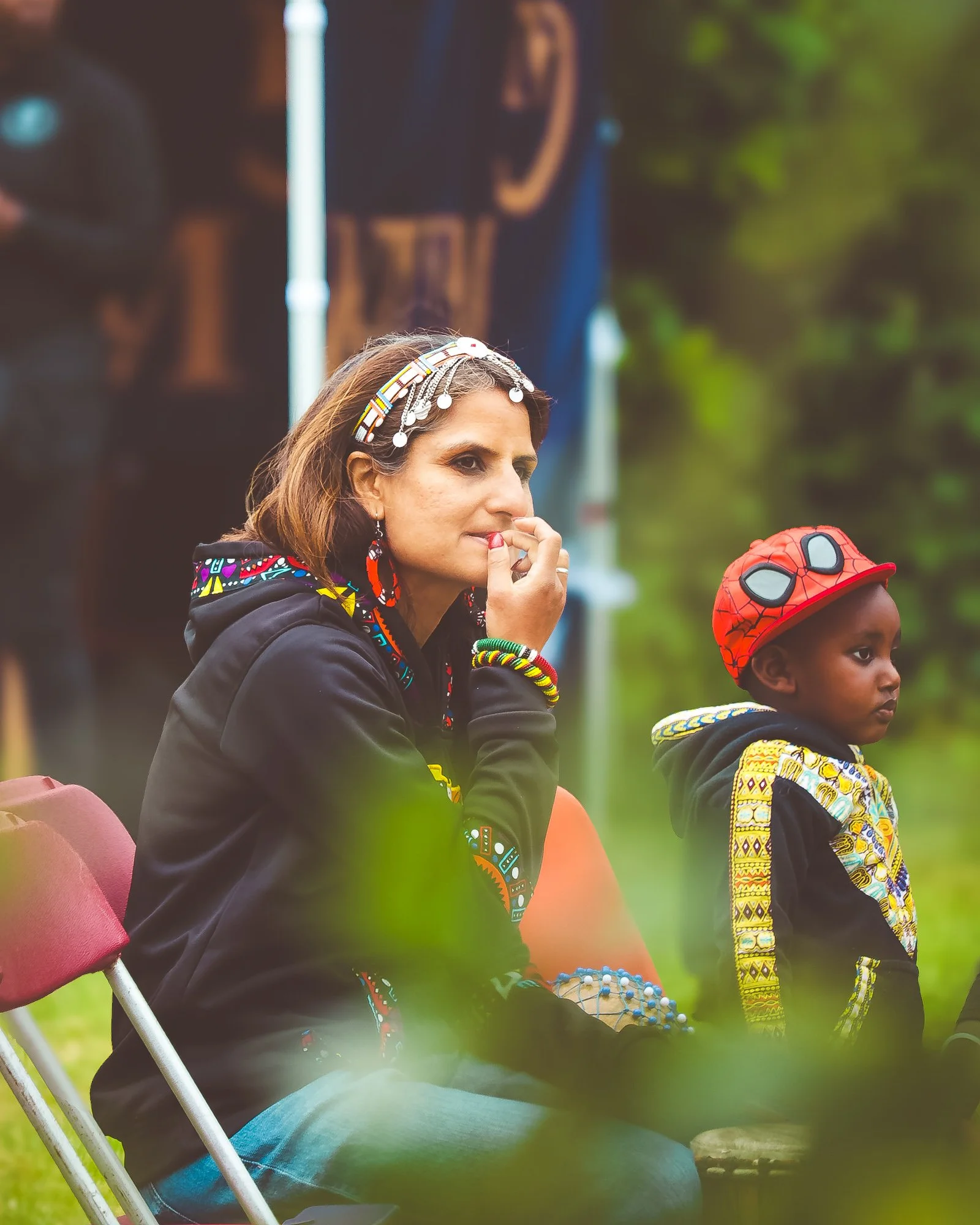 A woman and a young boy sitting outdoors, dressed in colorful clothing and accessories, with trees in the background.