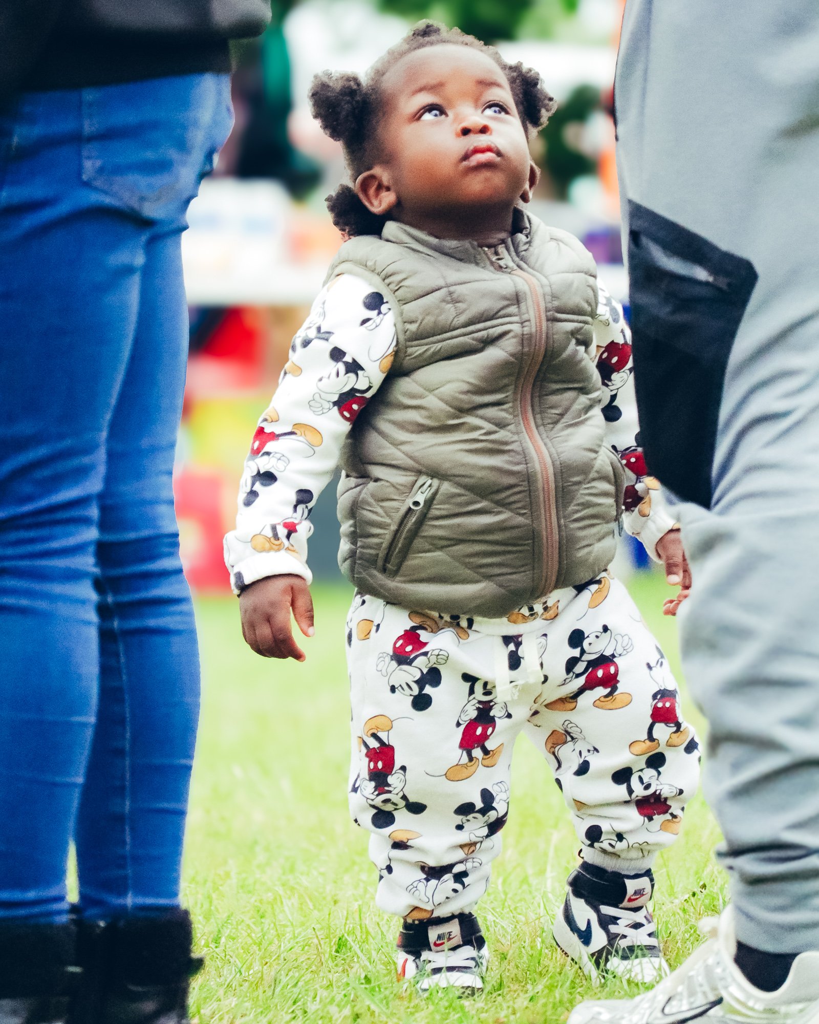 A young girl standing outdoors on grass, looking upward, surrounded by adults. She is wearing a Mickey Mouse-themed outfit, a beige vest, and sneakers.